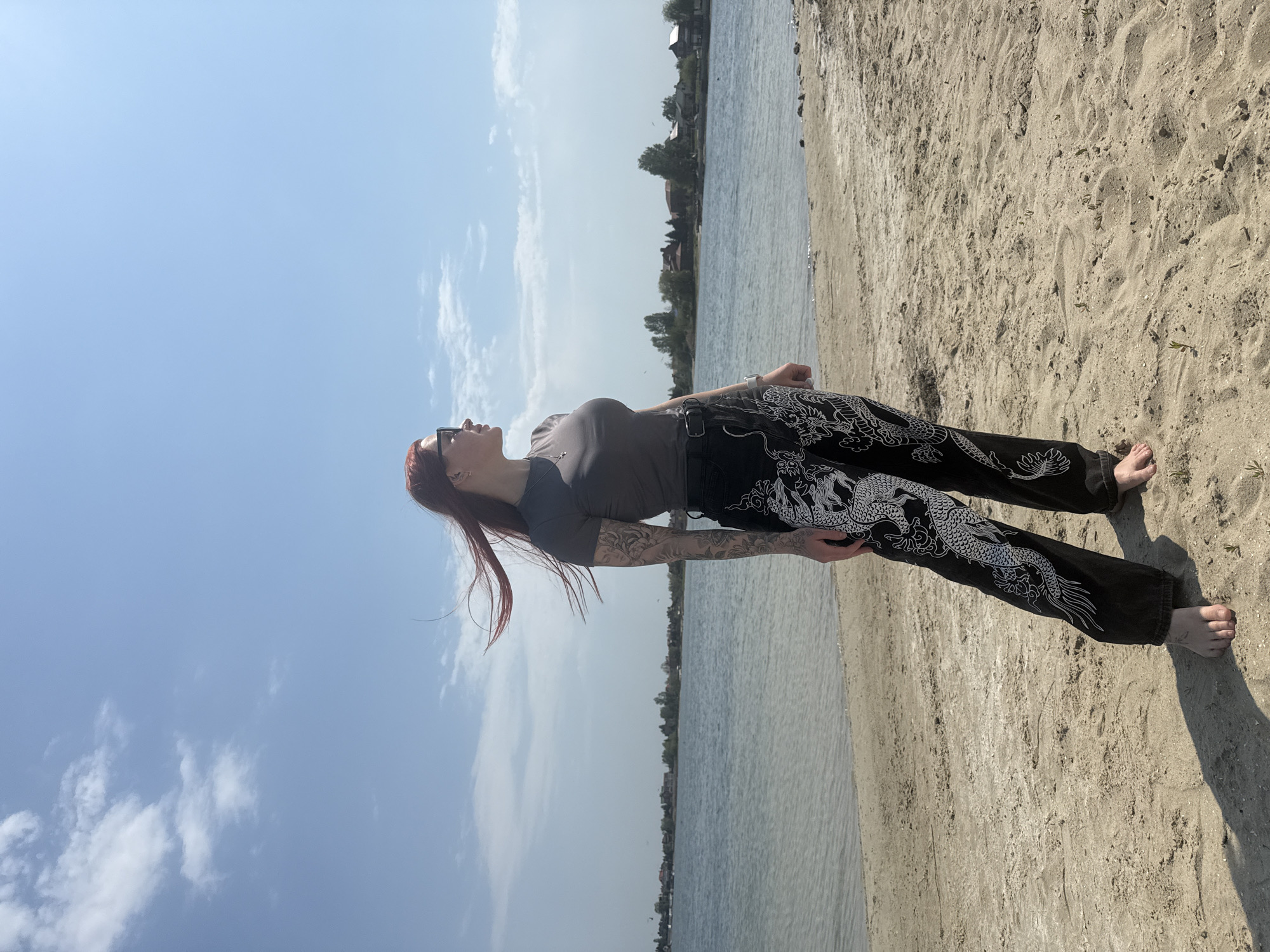 A woman with red hair stands barefoot on a sandy beach, looking up at the sky. She is wearing a dark-colored short-sleeved shirt and black pants with a white dragon design. The beach is bordered by a body of water and, in the distance, there are some buildings and trees, all under a partly cloudy sky. The location is in the city of Unknown, Unknown.