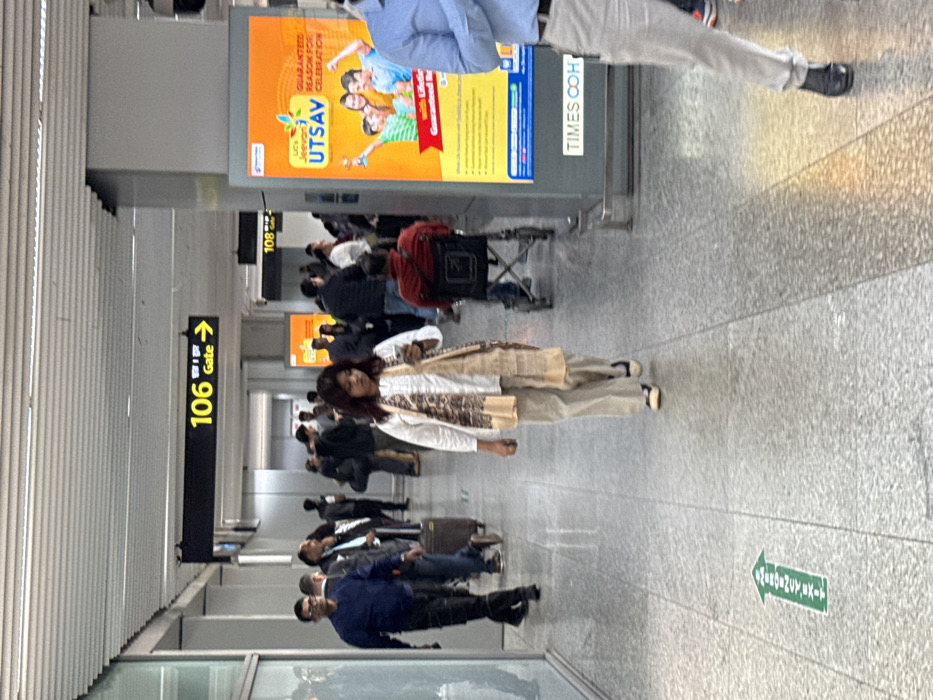 An airport hallway scene includes travelers walking and standing. A woman in a white patterned top and beige pants is centered. Signs for gates 106 and 108 are visible. Advertisements are displayed overhead. The floor is tiled, marked with a green arrow indicating an emergency exit. A person in a blue shirt walks towards the left of the frame, and another person in a light blue shirt is towards the right of the frame.