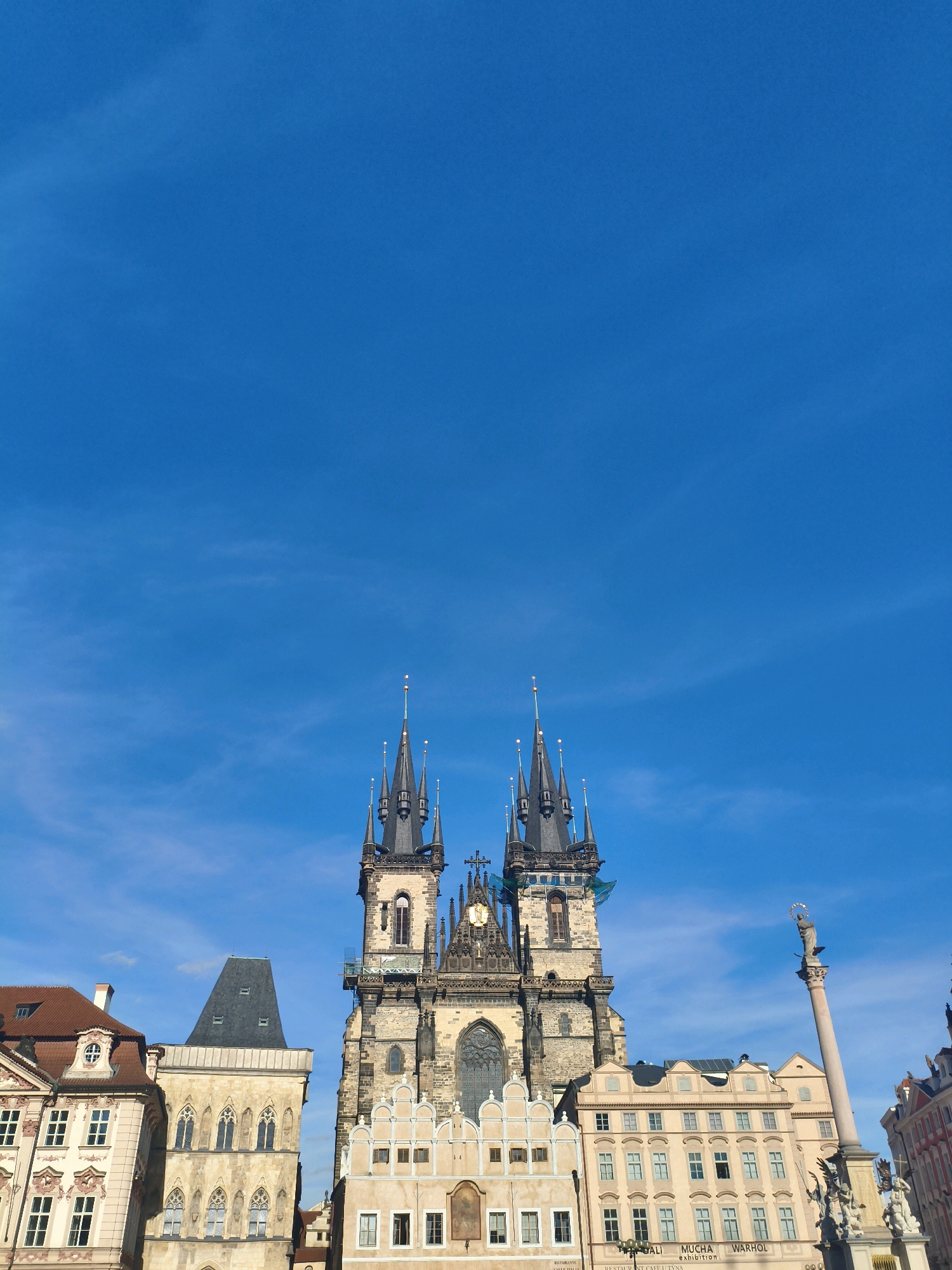 The image shows a wide shot of Old Town Square in Prague, Czechia, under a clear blue sky. Dominating the scene is the Church of Our Lady before Týn, a Gothic church with two tall, ornate spires. To the left of the church are several historic buildings with varied architectural styles, including a building with a red tiled roof and a distinctive pitched roof. To the right of the church is a tall column, likely the Marian column, topped with a statue. In front of the column, a monument with several statues is visible. The facades of the buildings display intricate detailing, windows, and decorative elements. A portion of the sky takes up the upper half of the frame, with faint wisps of clouds. The lower portion of the image is filled with the buildings and monuments characteristic of the square.
