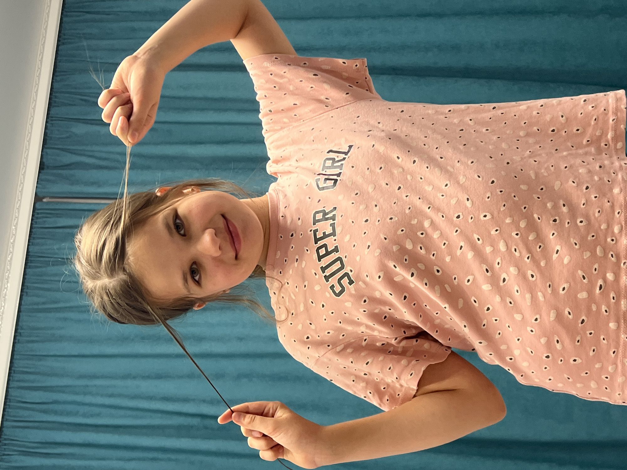 A young girl, approximately 7-9 years old, with light brown hair and a smiling expression, is captured in a close-up, chest-up shot. The image is taken from a slightly low angle, making her appear to be looking down at the camera. She is wearing a pink t-shirt with a white and black spotted pattern and the words "SUPER GIRL" printed in black letters on the front. Her right hand is raised, holding a few strands of her hair near the top, while her left hand is lower, holding a single, longer strand of her hair. The background consists of a teal or dark turquoise curtain, suggesting an indoor setting in Chaikovsky, Russia. The lighting is bright and even, indicating daytime. The girl's eyes are open and looking towards the viewer, and her lips are slightly parted in a gentle smile, conveying a playful and happy mood.