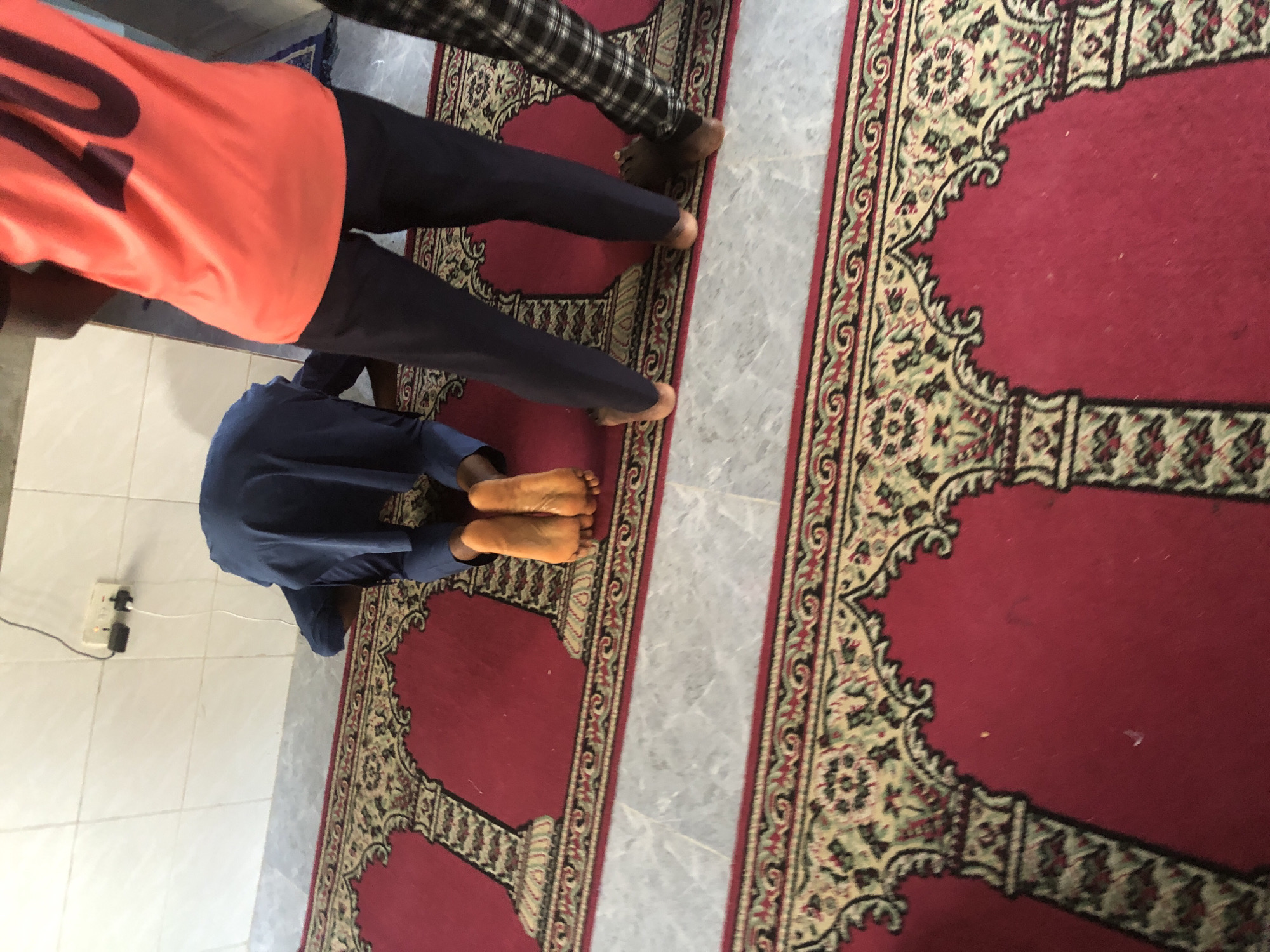 In Ado, Nigeria, an interior scene shows two individuals and a third person's legs. The foremost individual, wearing a dark blue garment, is in a prayer posture on a red carpet with intricate patterns. Their bare feet, stained orange, are visible, with the soles touching the carpet. The second individual, partially visible to the right, wears a bright orange t-shirt with the number '20' emblazoned on the back, and dark trousers. Their feet are bare. A third individual's legs, clad in plaid trousers and brown shoes, are positioned further to the right. The environment includes a tiled floor surrounding the carpet and a white tiled wall in the background, upon which an electrical outlet with a white cord is mounted.