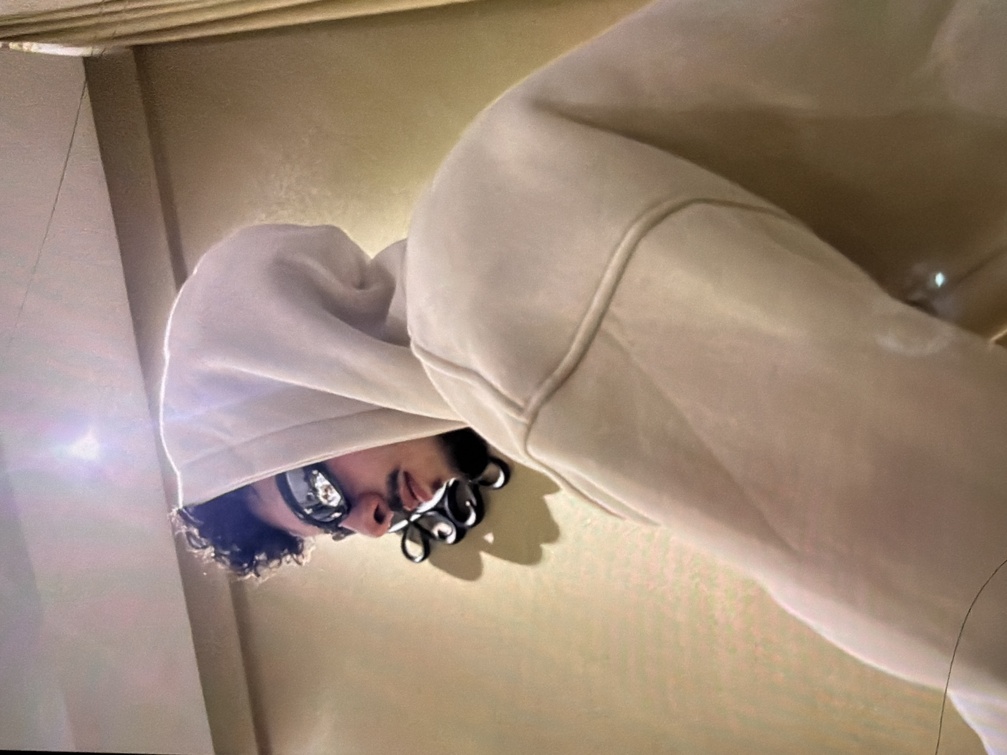 The image shows a person with curly hair wearing glasses and a white hooded sweatshirt. The person appears to be looking down at the camera. The background is a light-colored wall with a ceiling visible in the upper left corner. The location is unknown.