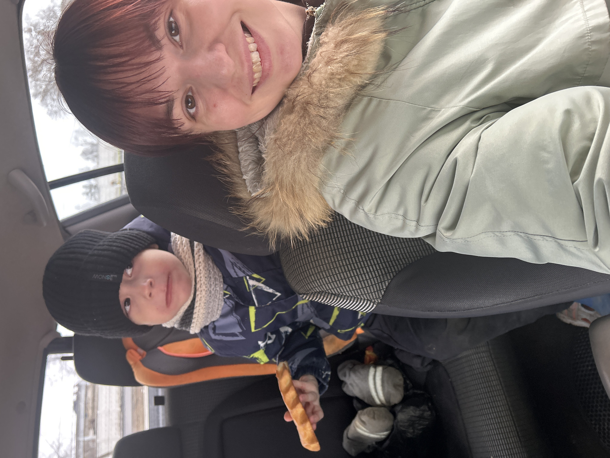 An adult woman takes a selfie inside a car, capturing herself and a young child in the backseat. The woman, with reddish-brown hair and dark eyes, smiles broadly at the camera. She wears a light green or sage-colored winter jacket with a prominent fur-trimmed hood. The child, seated in an orange and black car seat behind her, wears a black knitted beanie with the text "MONSI" and "SNOW" visible, a patterned blue winter jacket with neon yellow details, and a light-colored patterned scarf. The child holds a long, light brown, bread-like item, possibly a breadstick. Their expression is neutral and they look slightly upwards.
Through the car windows, a muted, overcast sky and bare trees or branches are visible, suggesting an outdoor, possibly wintry environment. Grey gloves are seen on the floor near the child's feet. The overall setting is the interior of a car during what appears to be a cold, cloudy daytime journey in Chaikovsky, Russia. The image conveys a moment of travel and casual interaction.