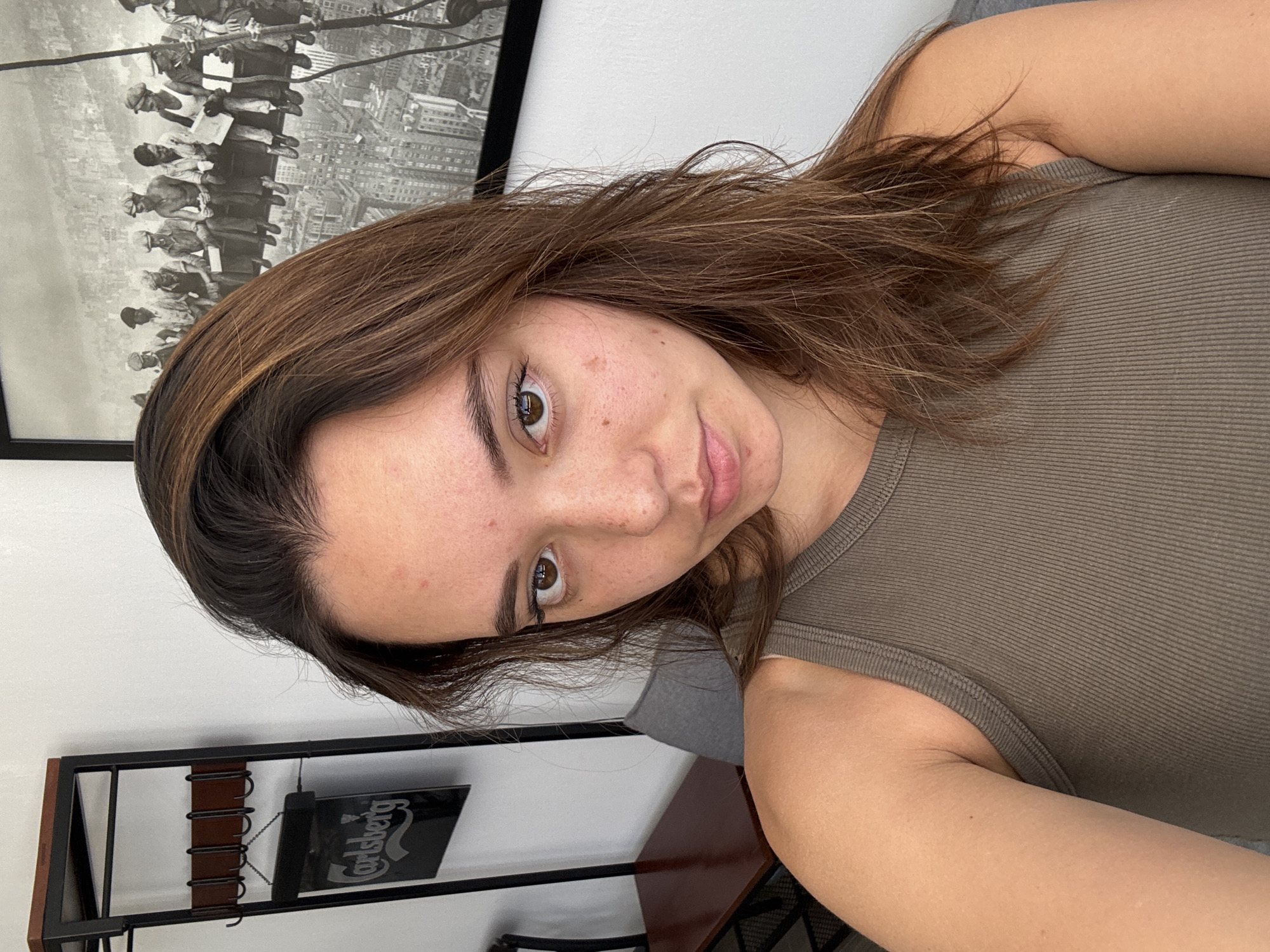 A young woman with brown hair and eyes is seen in a close-up, angled portrait. She is wearing a ribbed, olive-green tank top. In the background, there is a framed black and white photograph depicting men on a steel beam, seemingly constructing a building. To the left, a black metal shelf unit is visible with a dark, square item bearing a "Carlsberg" logo. The location, San Bernardino Tlaxcalancingo, Mexico, is unknown based on the visual elements of the image.