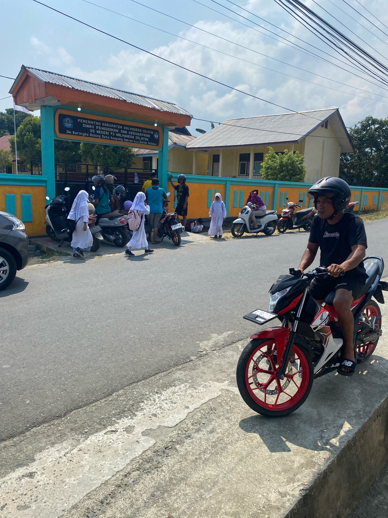 In Mandaong, Indonesia, on a sunny day, a young man wearing a black t-shirt, black shorts, a black helmet, and black sandals rides a red and white motorcycle. He is positioned on the right side of the frame, facing left, with his right leg extended. The motorcycle has a license plate attached to the front. Behind him, a yellow building with a corrugated metal roof is visible, bordered by a teal and orange fence. In front of the building is an arched gateway with signage that reads "PEMERINTAH KABUPATEN HALMAHERA SELATAN DINAS PENDIDIKAN DAN KEBUDAYAAN SD NEGERI 73 HALMAHERA SELATAN Jln. Barangka Dalong Desa Kampung Hakian Kec. Bacan Selatan".
Several other people are present. Two young girls in white hijabs and dresses are walking away from the viewer towards the gateway. Another girl in a white hijab is seen from the side, walking behind the first two. A woman in a purple hijab and patterned shirt is seated on a white scooter. A man in a blue t-shirt stands near a motorcycle, with his arm raised. Another man in a black t-shirt stands with his back to the viewer. Various parked motorcycles are visible in front of the fence, along with a dark grey car on the far left of the image. The scene appears to be a street outside of a school. The road is asphalt, with a concrete curb and sidewalk. The sky is bright blue with scattered clouds.