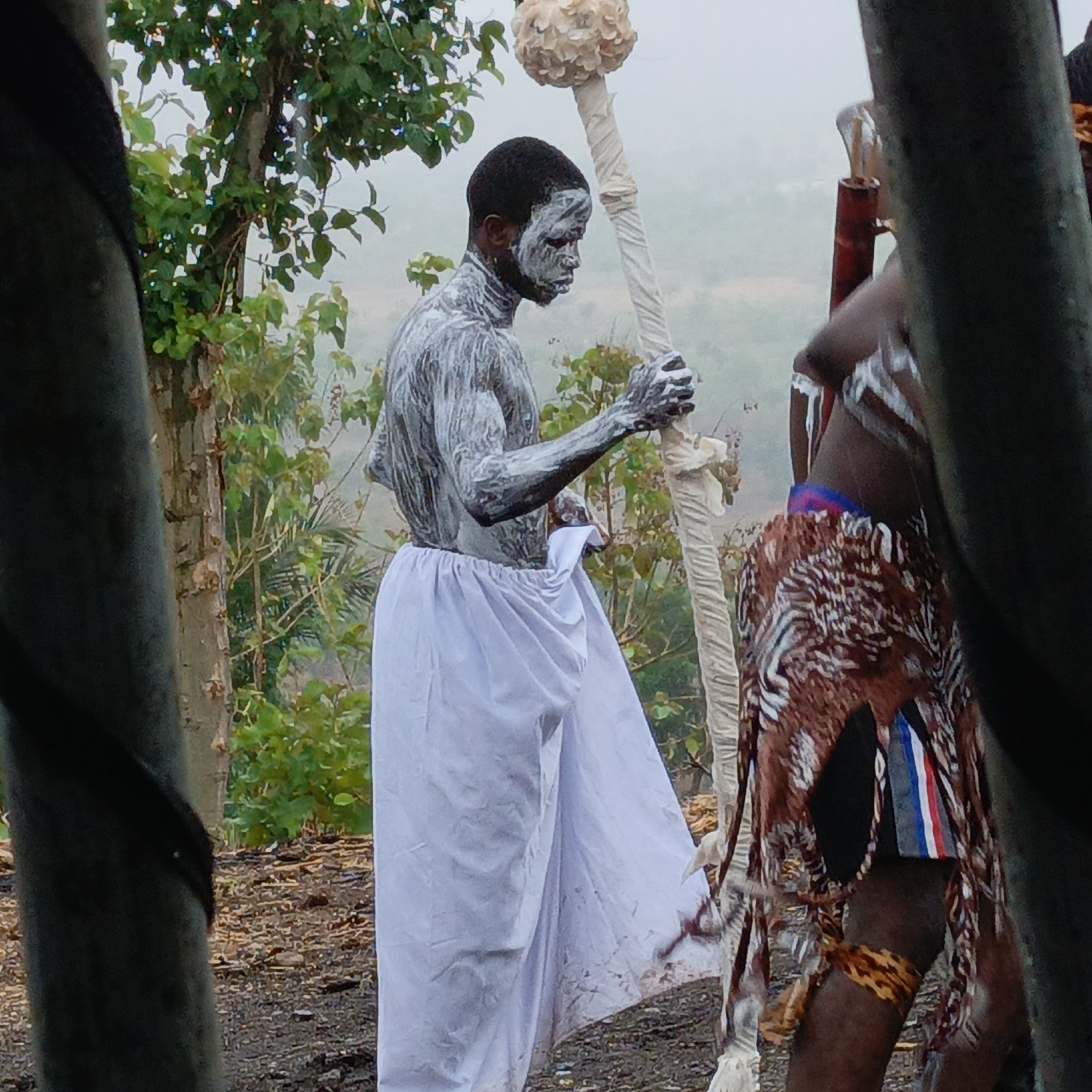In a location identified as Boher, Nigeria, the image depicts two individuals engaged in what appears to be a cultural or ritualistic activity. One man, prominently positioned, is painted with white pigment covering his face and torso. He wears a long, white garment and holds a tall staff wrapped in white material, topped with a round, organic-looking object. The second man is partially visible, his lower body adorned with a patterned cloth skirt. He stands near the first man. They are in an open area, possibly a clearing or at the edge of a woodland, with trees and foliage visible in the background. The weather appears overcast, casting a muted light over the scene. A man is standing behind trees in the forefront of the image.