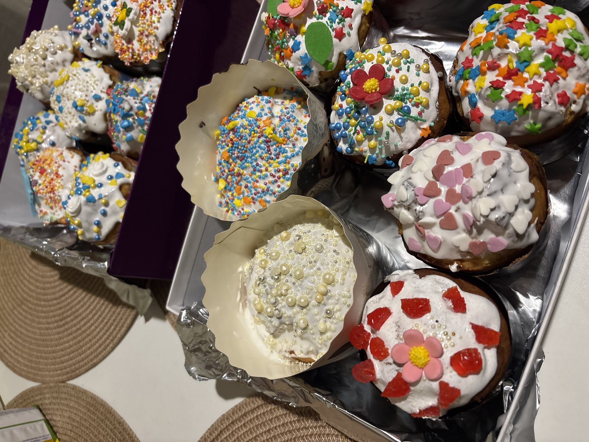 The image shows two containers of decorated Easter cakes. The container on the left is a dark purple box holding six cakes with white icing and various colorful sprinkles. The container on the right is a shallow tray lined with aluminum foil holding six more Easter cakes. These cakes are individually wrapped in paper cups and decorated with white icing, sprinkles, and candied fruit. The scene appears to be indoors on a table with a placemat.
There is no indication that this image was taken in Pokotylivka, Ukraine.