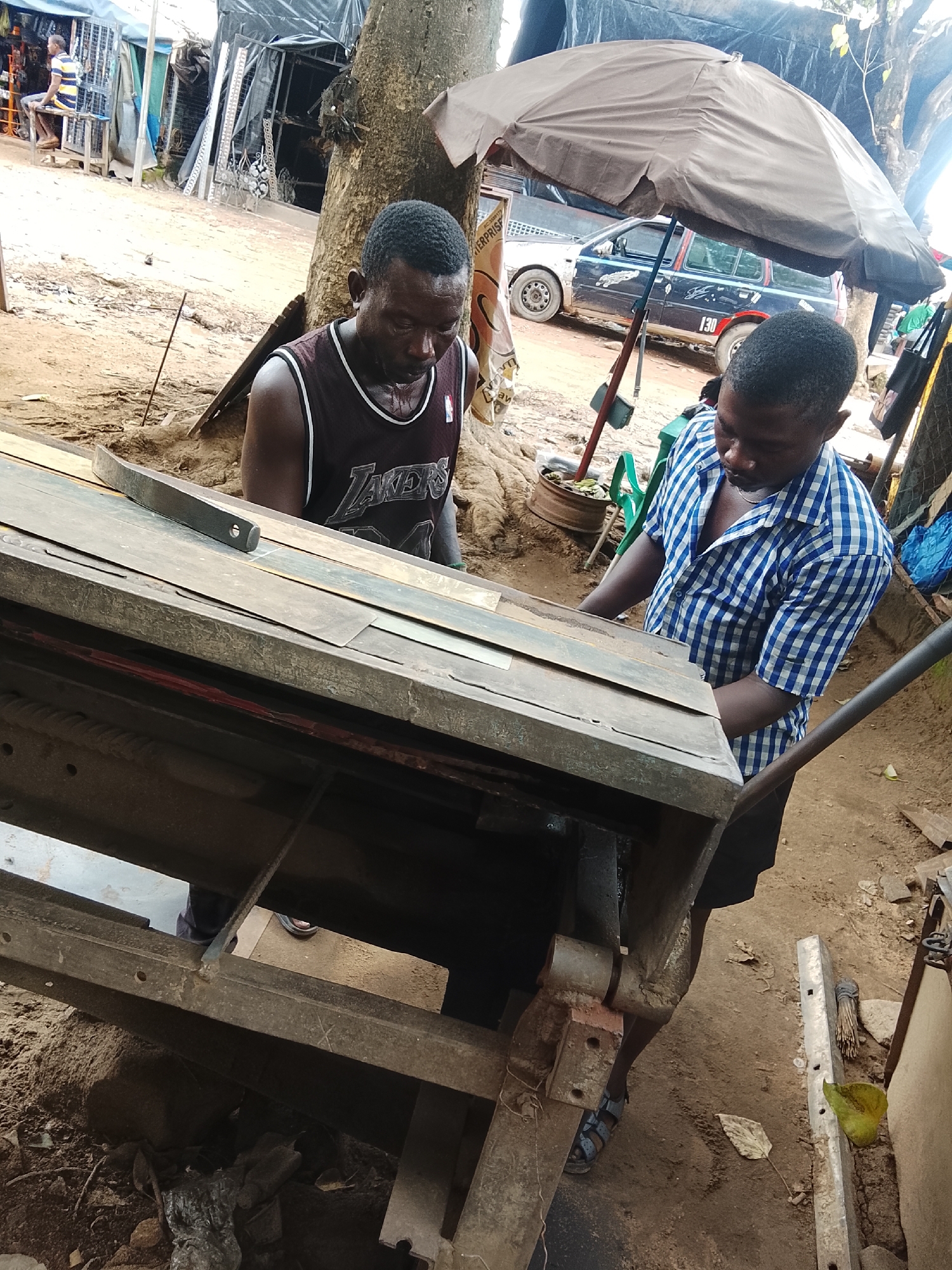 The image depicts an outdoor scene in what appears to be a workshop or market stall in Abuja, Nigeria. Two men are actively engaged with a piece of machinery, possibly a metal cutting or shaping device, which is made of heavy, weathered metal and wood. The man in the foreground, wearing a blue and white checkered shirt, is focused on the machinery, with his hands positioned near it. He is wearing sandals. Behind him, another man, wearing a dark tank top with "Lakers" printed on it, also seems to be involved with the task, looking down at the equipment. The ground is dirt and sand, with debris scattered around, including leaves and what appears to be discarded materials. In the background, there are structures covered with tarpaulin, a parked car with visible license plates and stickers, and an open umbrella providing shade. The overall impression is a working environment in a somewhat dusty and informal setting.