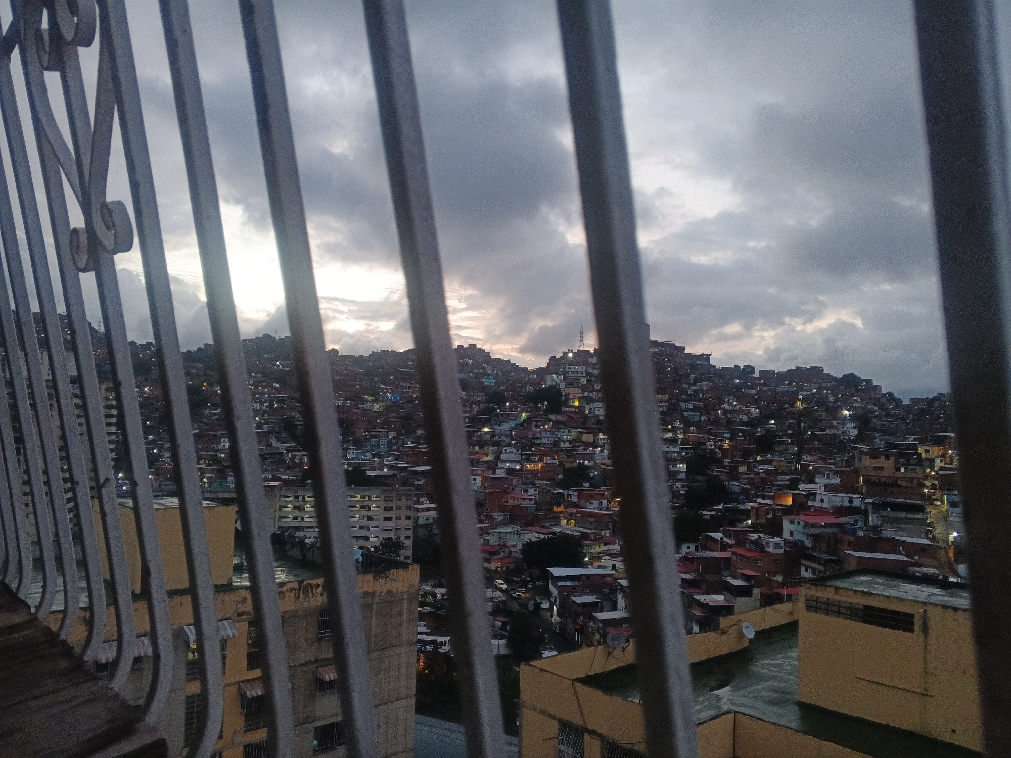 The image presents a wide-angle view of a densely populated urban landscape, likely from a high vantage point such as a balcony, framed by vertical metal railings in the foreground. The sky is overcast and cloudy, with patches of brighter light visible on the horizon, suggesting either dawn or dusk.
In the foreground, a gray metal railing with decorative scrollwork on the left side partially obscures the view. Beyond the railing, the scene unfolds into a vast expanse of buildings covering multiple hillsides. These buildings appear to be a mix of informal settlements or "barrios," characterized by their close proximity, varying sizes, and irregular construction, alongside some larger, multi-story structures. Many houses are painted in different colors, and their flat roofs are visible, some with small satellite dishes or water tanks.
As the light fades or begins to emerge, scattered artificial lights from windows and streetlights are visible across the cityscape, casting a warm glow against the muted tones of the sky and buildings. A prominent electricity pylon or transmission tower can be seen on a distant hilltop against the horizon. The overall impression is one of a vast, sprawling city waking up or settling down, with a sense of urban density and an intimate view of the residential areas built into the natural terrain. No specific activities are taking place, and there is no visible text in the image. The setting, as suggested by the context, is characteristic of Caracas, Venezuela.