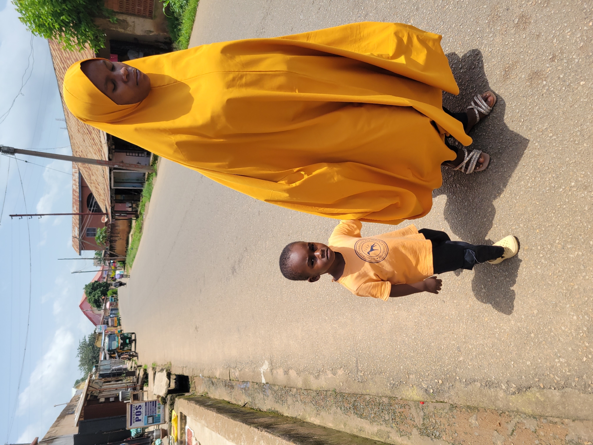 In Jos, Nigeria, a woman in a vibrant yellow hijab walks alongside a young boy on a paved street. The woman's hijab drapes over her, obscuring most of her body, and she wears decorative sandals. The boy, wearing an orange t-shirt with a circular emblem and dark pants, walks to her left, holding her hand. Their shadows stretch across the asphalt road, indicating a sunny day. The street is lined with buildings, some appearing to be shops with signs visible, suggesting a commercial area. A sign for "MONIEPOINT POS" is prominent on the left side of the street. Several motorized tricycles, commonly known as "keke napep" in Nigeria, are parked or moving in the background, along with other pedestrians and some vegetation. The sky above is partly cloudy.