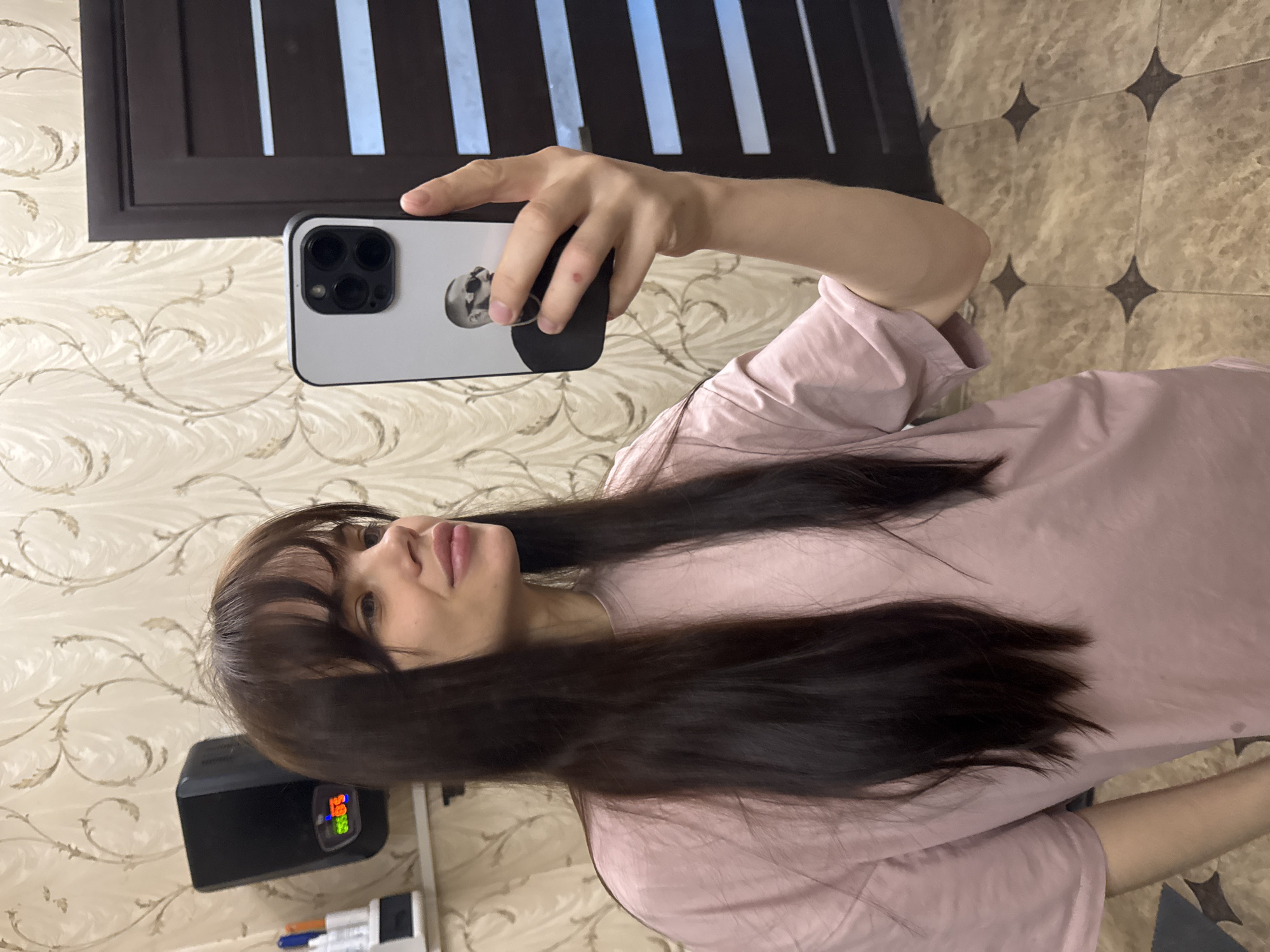 A young woman with long, dark brown hair and bangs is taking a selfie with her iPhone. She is wearing a light pink t-shirt. The phone has a white case with a graphic on the back. The background includes a wall with floral patterned wallpaper, a dark brown door with vertical panels, and a floor with a geometric pattern. To the lower left, there is a black object with a digital display. The location in Rostov-on-Don, Russia, cannot be determined from the image.