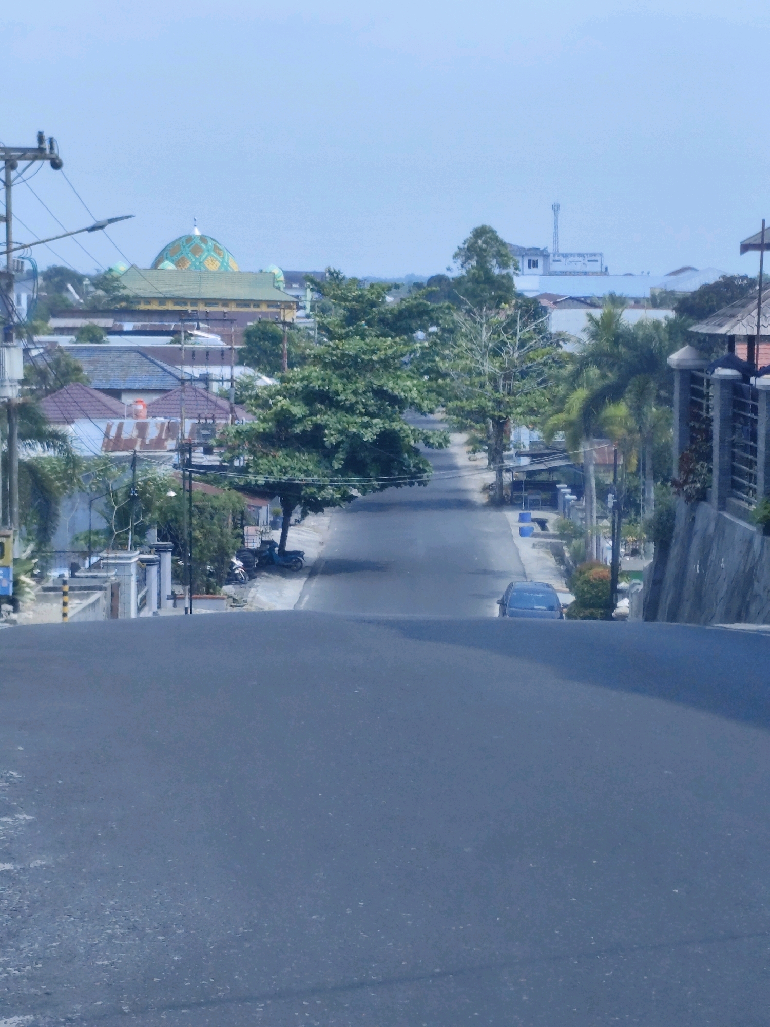 The image shows a street in Pangkalan Bun, Indonesia, sloping downwards. On the left side of the street, there are houses with various roofing materials, including corrugated metal and tiles. A mosque with a prominent green and gold dome is visible in the distance. There are also utility poles and trees lining the street. On the right side of the street, there are more houses, some with concrete walls and tiled roofs, and some vegetation. A gray car is parked on the street. A motorcycle is parked on the left side of the street, near some houses. The sky is overcast.