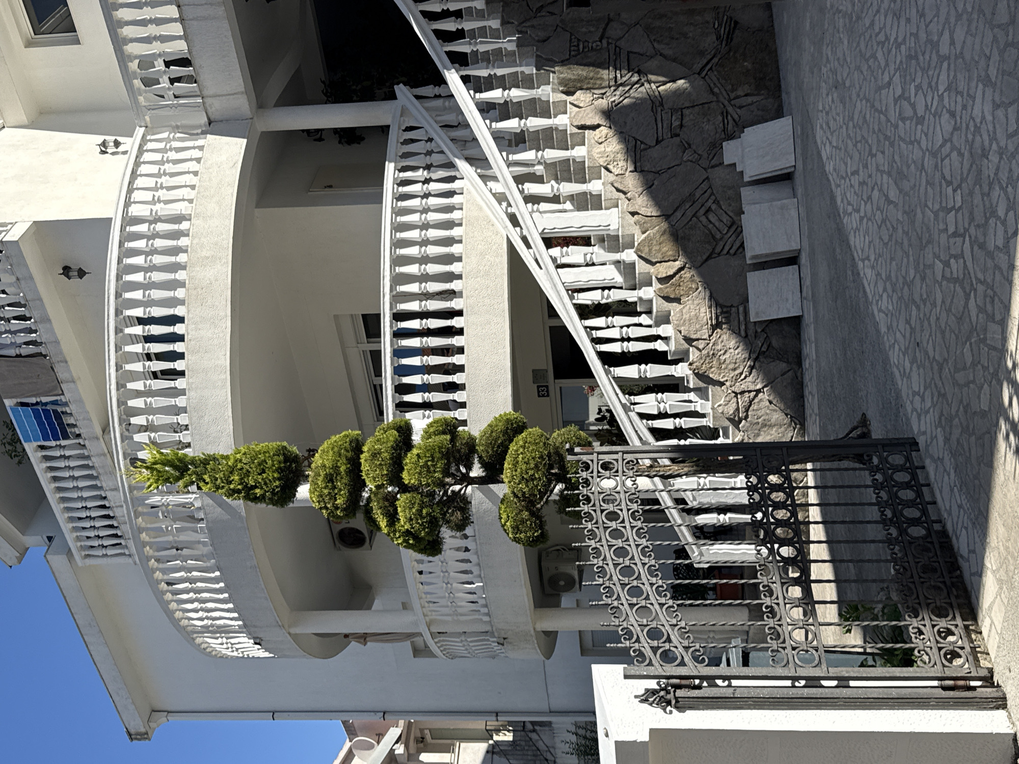 The image shows a white building with a curved balcony, white railings, and white stairs leading up to the building's entrance. A decorative tree is in the foreground, to the left of the stairs and in front of a black metal gate. The building appears to be a residential property or a small hotel. In the background, there is a clear blue sky. The scene is likely located in Budva, Montenegro, a popular tourist destination known for its coastal scenery and architecture.
