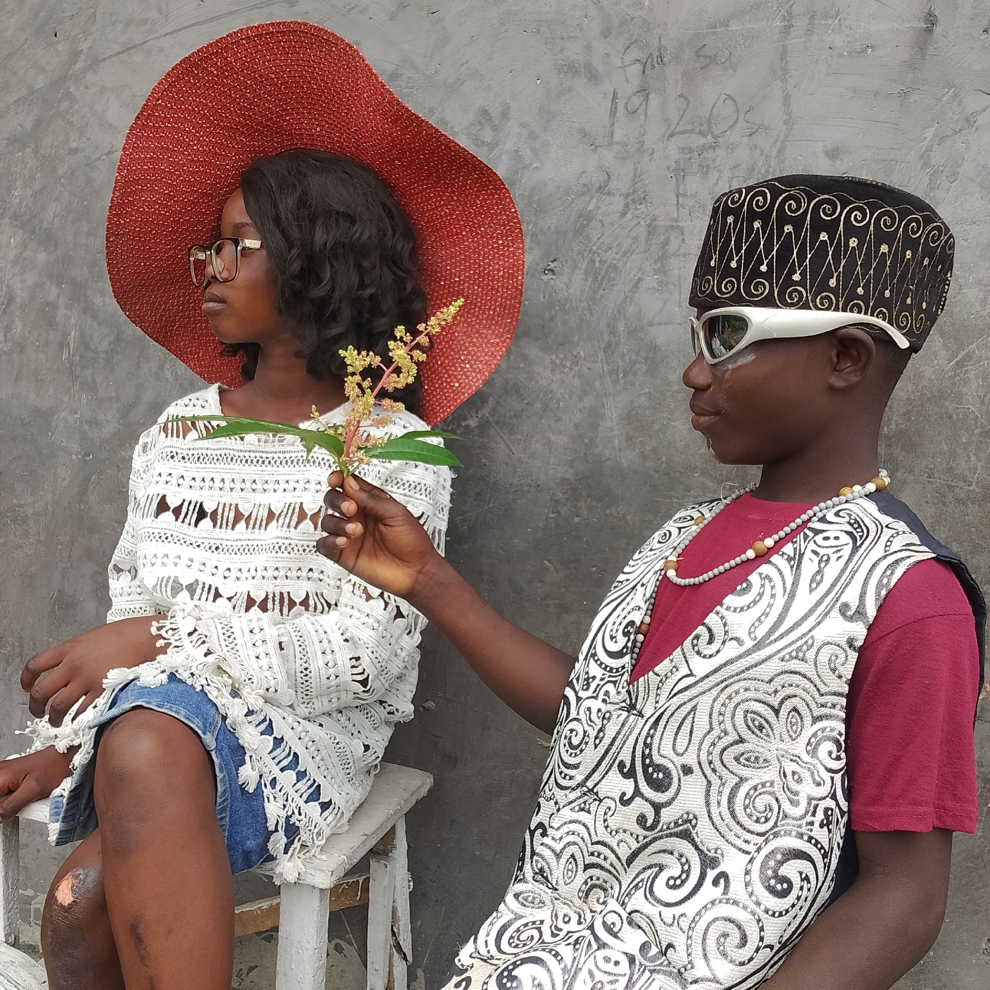 In Ado, Nigeria, two children are positioned against a textured, gray concrete wall. The child on the left, a girl, wears a wide-brimmed red hat and clear-rimmed glasses. She has dark, wavy hair and is dressed in a white, crocheted top with intricate patterns and denim shorts. Her left arm rests on her lap, and her right hand holds a small branch with yellow flowers. The child on the right, a boy, wears a black, embroidered cap with gold swirling patterns and futuristic-looking white sunglasses. He is dressed in a maroon t-shirt, over which he wears a black and white patterned vest. A beaded necklace, consisting of gray and wooden beads, adorns his neck. The boy's right arm is extended, holding the flower branch towards the girl. The overall scene appears to be an outdoor portrait.