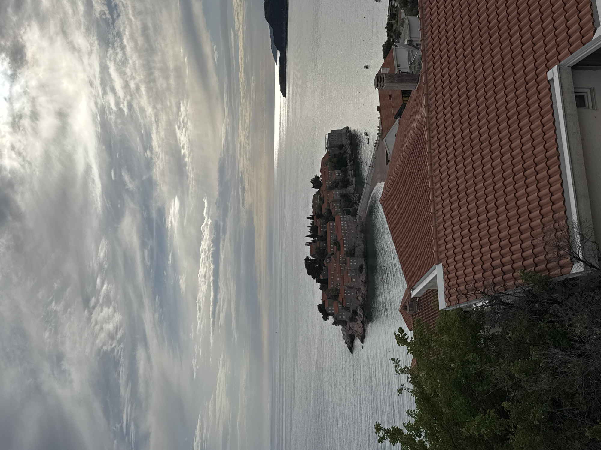 The image captures a landscape in Sveti Stefan, Montenegro, featuring a small island with closely-packed buildings and terracotta roofs. The island is connected to the mainland by a narrow isthmus. The Adriatic Sea is calm and reflects the sky. In the foreground, the roof of a building with terracotta tiles and a green tree are visible, suggesting a vantage point from a nearby structure. The sky is filled with elongated, layered clouds, with the sun’s reflection on the water. A distant landmass is present on the horizon.