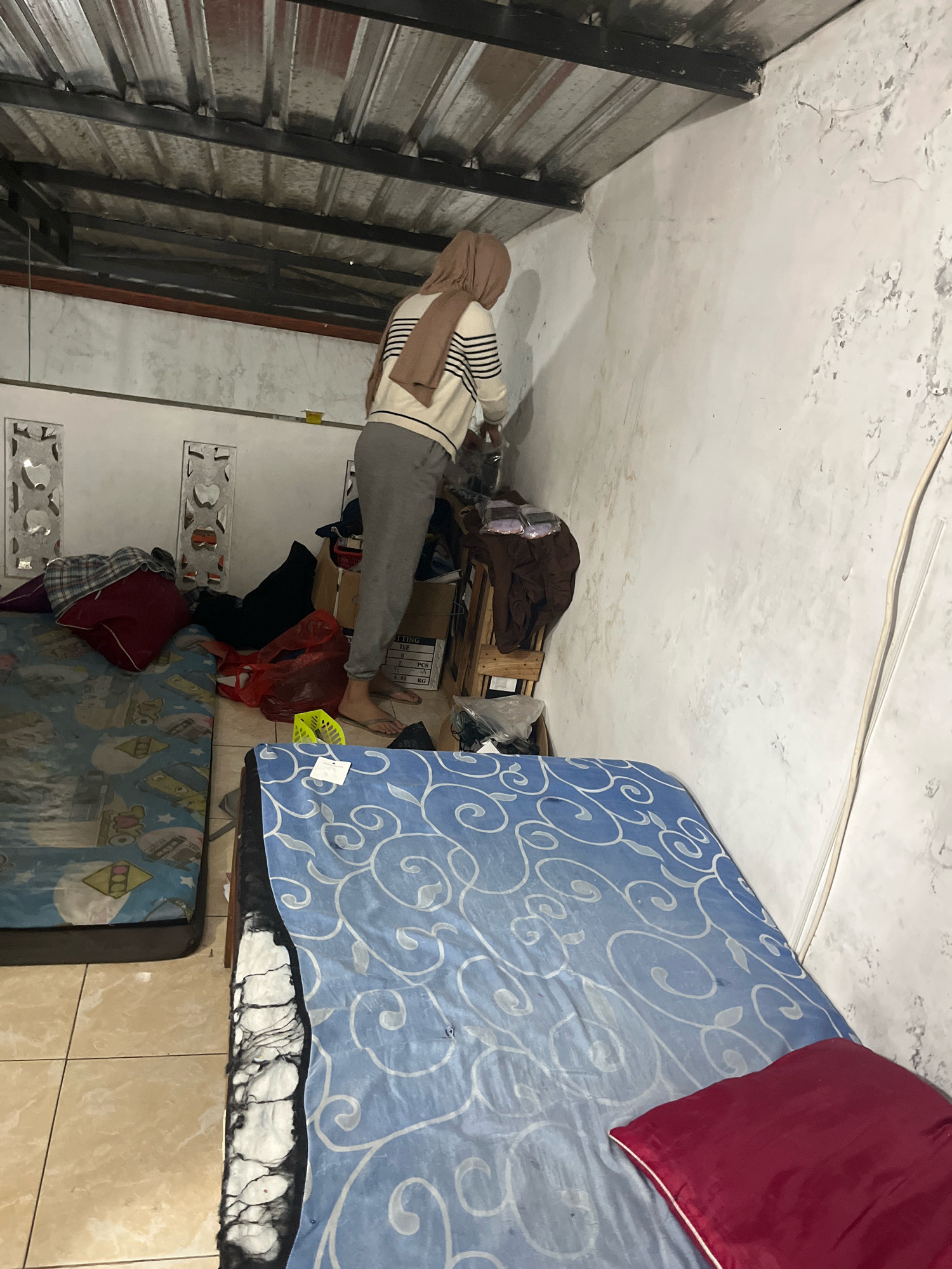 The image depicts a cluttered room, possibly a temporary shelter or living space, in Matesih, Indonesia. The room features two mattresses on the floor. One mattress, on the left, is covered with a blue sheet patterned with cartoon characters. The other mattress, in the foreground, is covered with a blue sheet featuring a white swirling design and a red pillow. In the background, a person wearing a hijab and a striped sweater is standing on a makeshift ladder, appearing to be reaching for something on a shelf or wall. The walls are white and show signs of wear and tear, with peeling paint and water stains. The ceiling is made of corrugated metal. Various items, including bags, boxes, and clothing, are scattered around the room, contributing to the overall sense of disarray. The lighting suggests it is nighttime or the room is dimly lit.