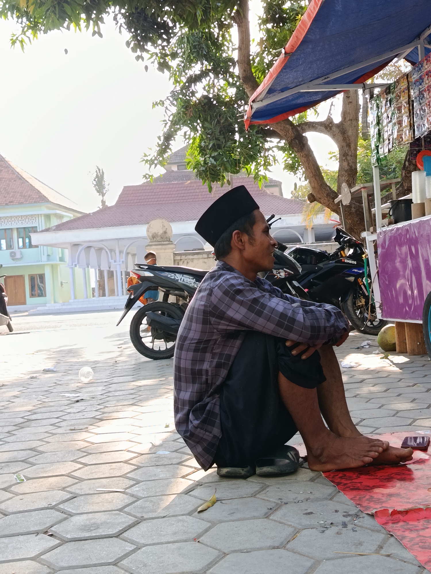 A middle-aged Indonesian man wearing a black peci and a grey and black checkered long-sleeved shirt sits on the ground, facing right, with his legs crossed and his hands resting on his knees. He is barefoot, wearing black slippers. He is positioned on a hexagonal brick pathway in front of a stall covered by a blue and red tarp. Behind him, several motorcycles are parked. In the background, a building with a red tiled roof and white arched colonnades is visible, along with a light green building. The scene appears to be outdoors, possibly in a public area or market, with trees and bright sunlight contributing to the environment. The location is identified as Ramban Wetan, Indonesia.