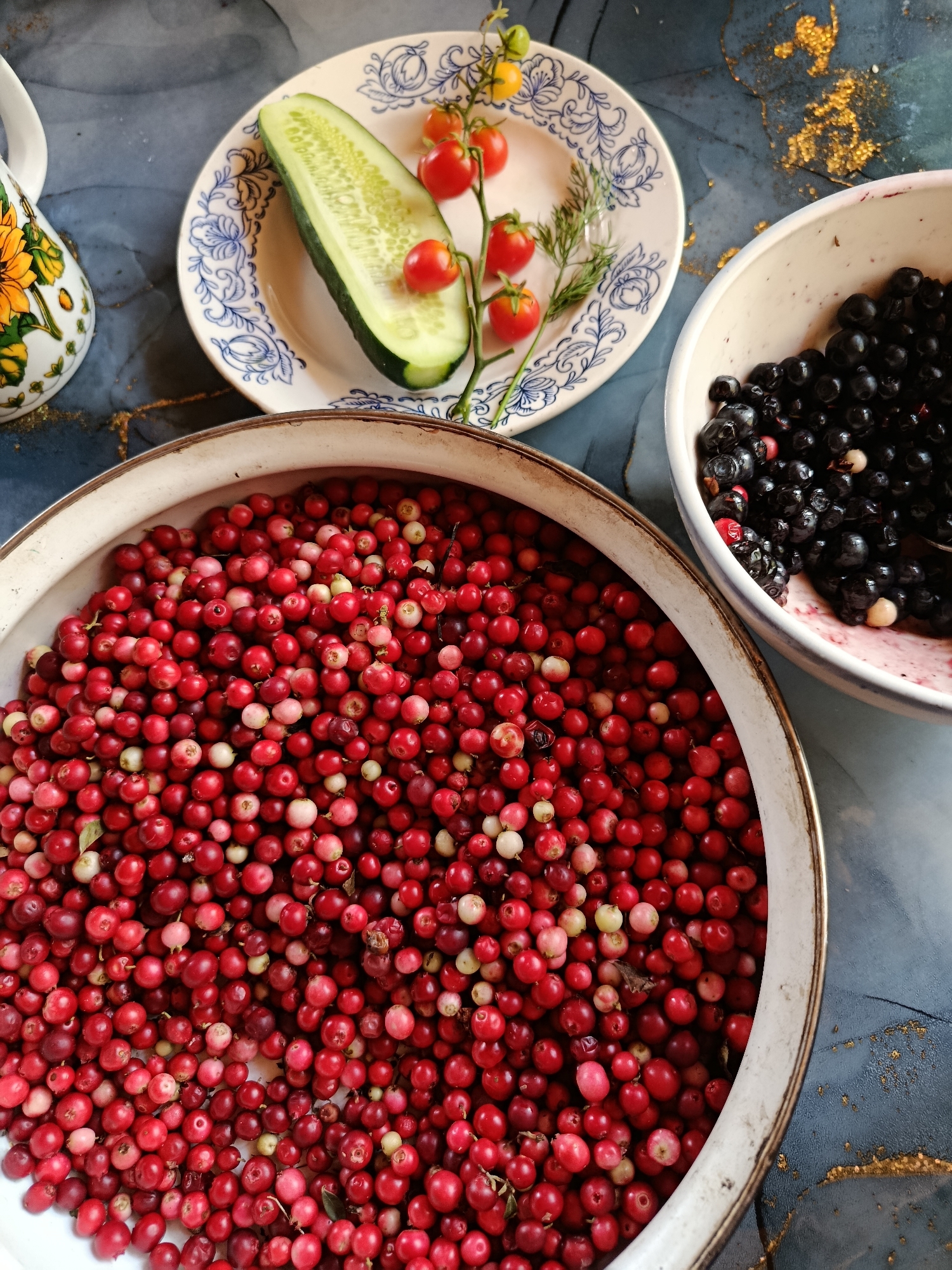 The image displays a collection of fresh berries and vegetables arranged on a table with a dark blue, textured surface with gold flecks. In the foreground, a large, shallow metal bowl is filled with a mound of vibrant red lingonberries. To the upper right of the lingonberries, a white ceramic bowl contains dark blue blueberries. In the upper center of the image, a small white plate with blue decorative trim holds a sliced cucumber and a sprig of cherry tomatoes with a sprig of dill. To the left of the plate, a portion of a white ceramic pitcher with a sunflower design is visible. The scene appears to be a still life composition of freshly harvested produce, likely intended for culinary use. There is no indication of location in the city of Vereya, Russia.