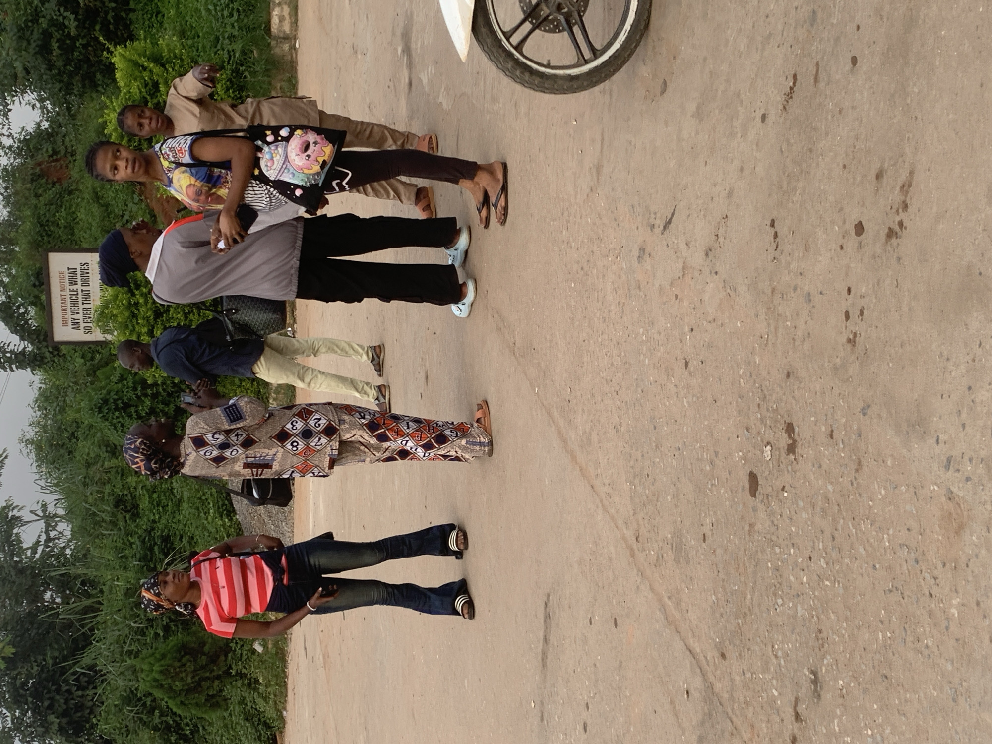 In Shagamu, Nigeria, a group of six individuals stands on a paved, concrete surface. A woman on the left wears a pink and white striped t-shirt, dark jeans, and sandals. Beside her, another woman is dressed in a patterned top and pants, with sandals. A man in a dark shirt and light pants stands to the right of them. Next, a woman in a grey hooded jacket and black pants, wearing a dark headscarf, is interacting with another woman wearing a colorful t-shirt adorned with images and carrying a tote bag. The last woman, on the far right, is in a tan outfit and sandals. A white motorcycle is partially visible in the lower right corner. Behind the people, lush green vegetation and trees line the background. A sign with important notice text is mounted on a pole. The ground is a textured concrete surface with visible stains and debris.