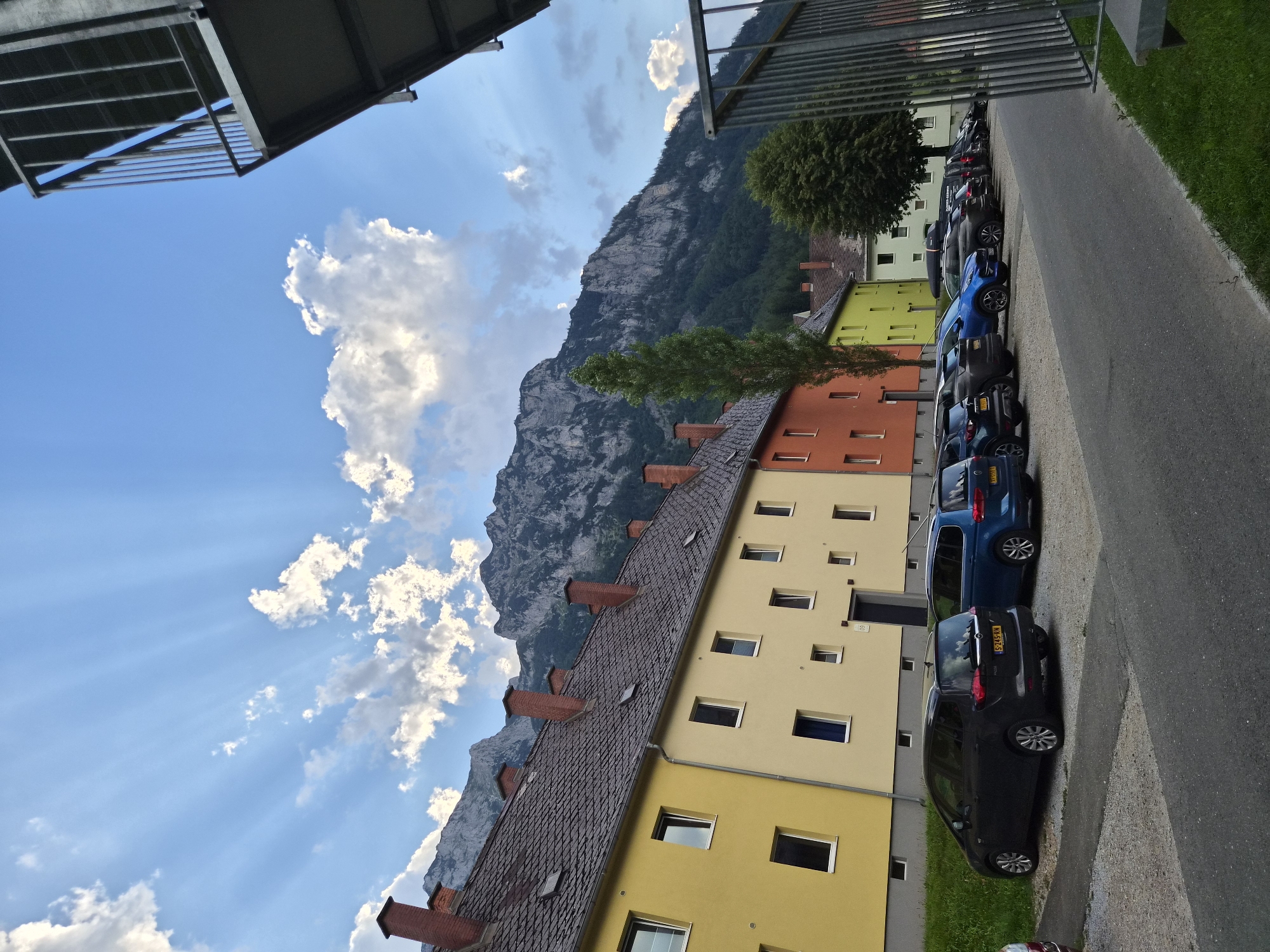 The image shows a view of a residential building in Eisenerz, Austria, with a mountain range in the background under a partly cloudy sky with crepuscular rays. The building has a facade with sections painted in yellow, light yellow, and orange. Several cars are parked in a lot in front of the building. The parking lot is paved with gravel and asphalt. A metal railing is visible in the foreground on the right side of the image. The mountain range is covered in trees and rocky outcrops. The sky is blue with scattered white clouds.