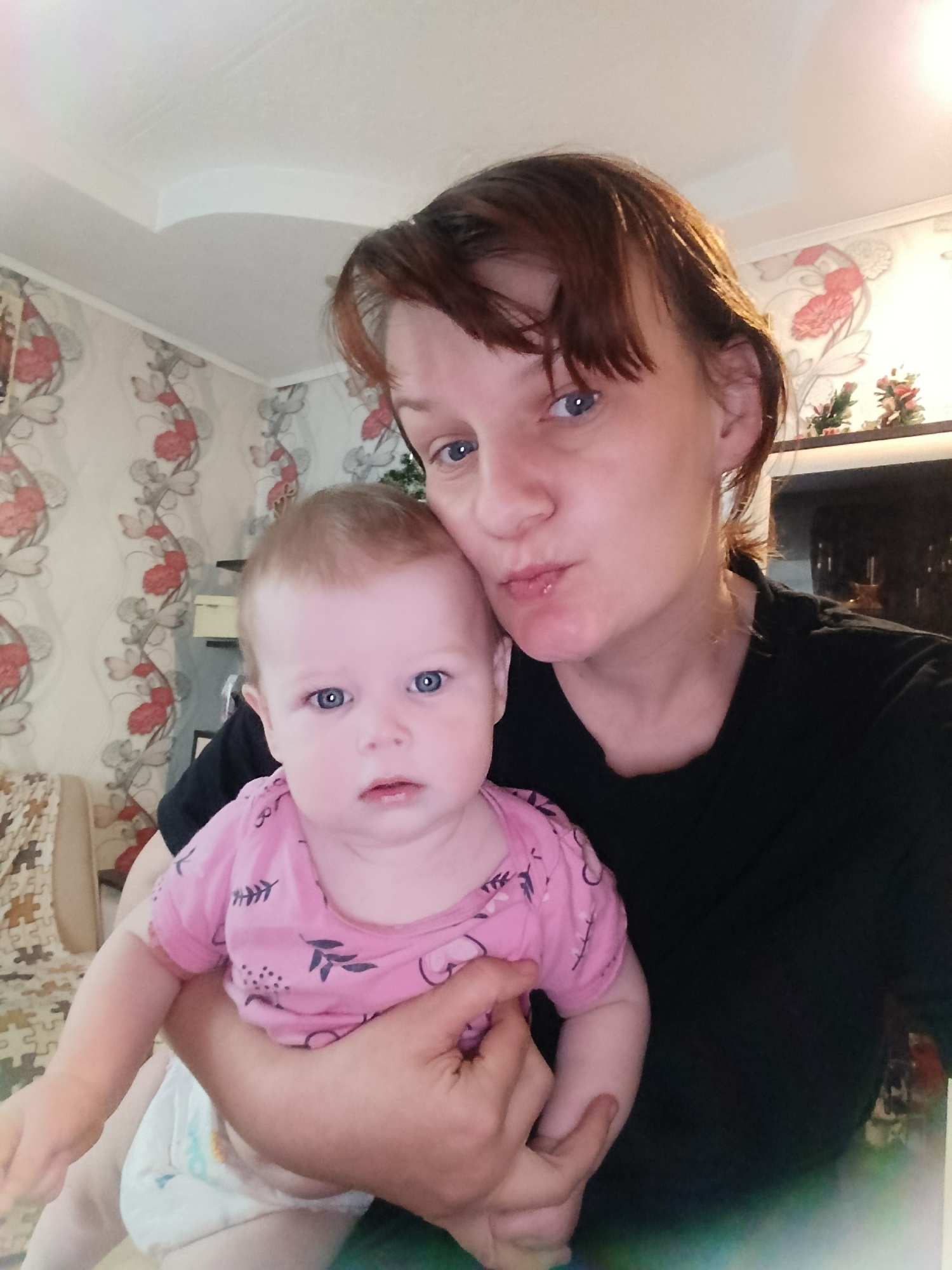 A photograph depicts a fair-skinned woman holding a baby, both with light blue eyes, in an indoor environment. The woman has short brown hair with bangs and is wearing a black t-shirt. Her face is close to the baby's, and she is puckering her lips. The baby is wearing a pink t-shirt with a pattern of small black branches and a diaper. The background includes floral wallpaper, a ceiling with decorative molding, a shelf with decorative items, a piece of furniture covered with a puzzle-like pattern, and a dark object that could be a television. The location appears to be an interior living space in a city that is not known.