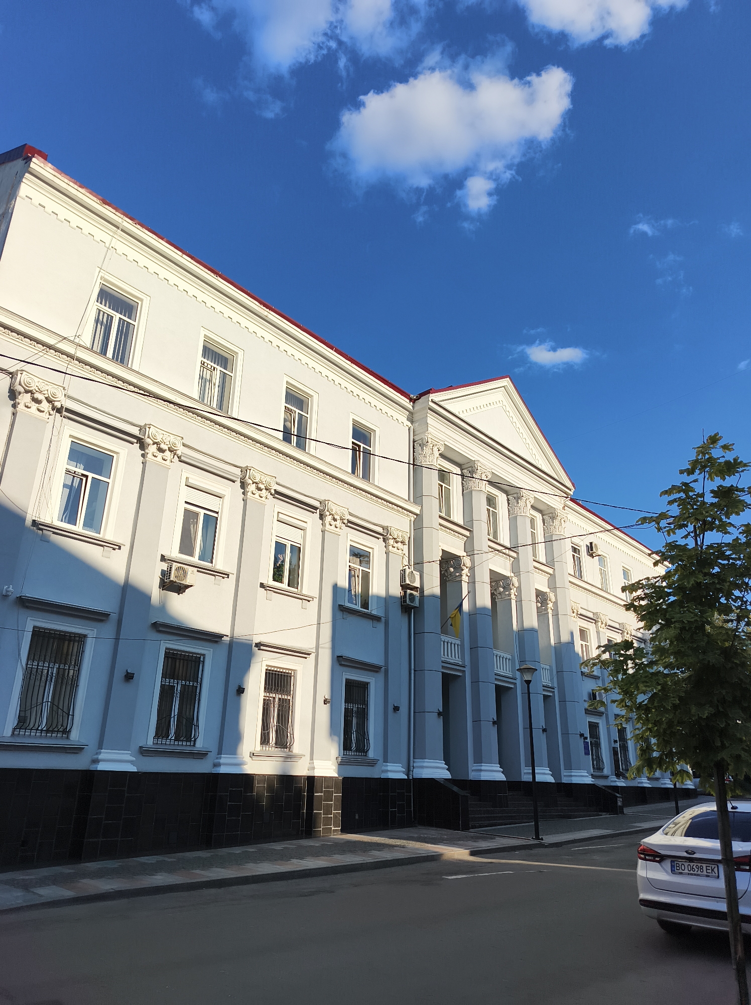 A large, light blue building with a red roof and white decorative elements stands prominently under a bright blue sky with scattered white clouds. The building features multiple windows, some with decorative grates, and a portico with classical columns. A street runs in front of the building, with a white car parked on the right side. The scene suggests a daytime view in an urban setting. The building is located at 2 Tantsiova Street, Ternopil, Ukraine.