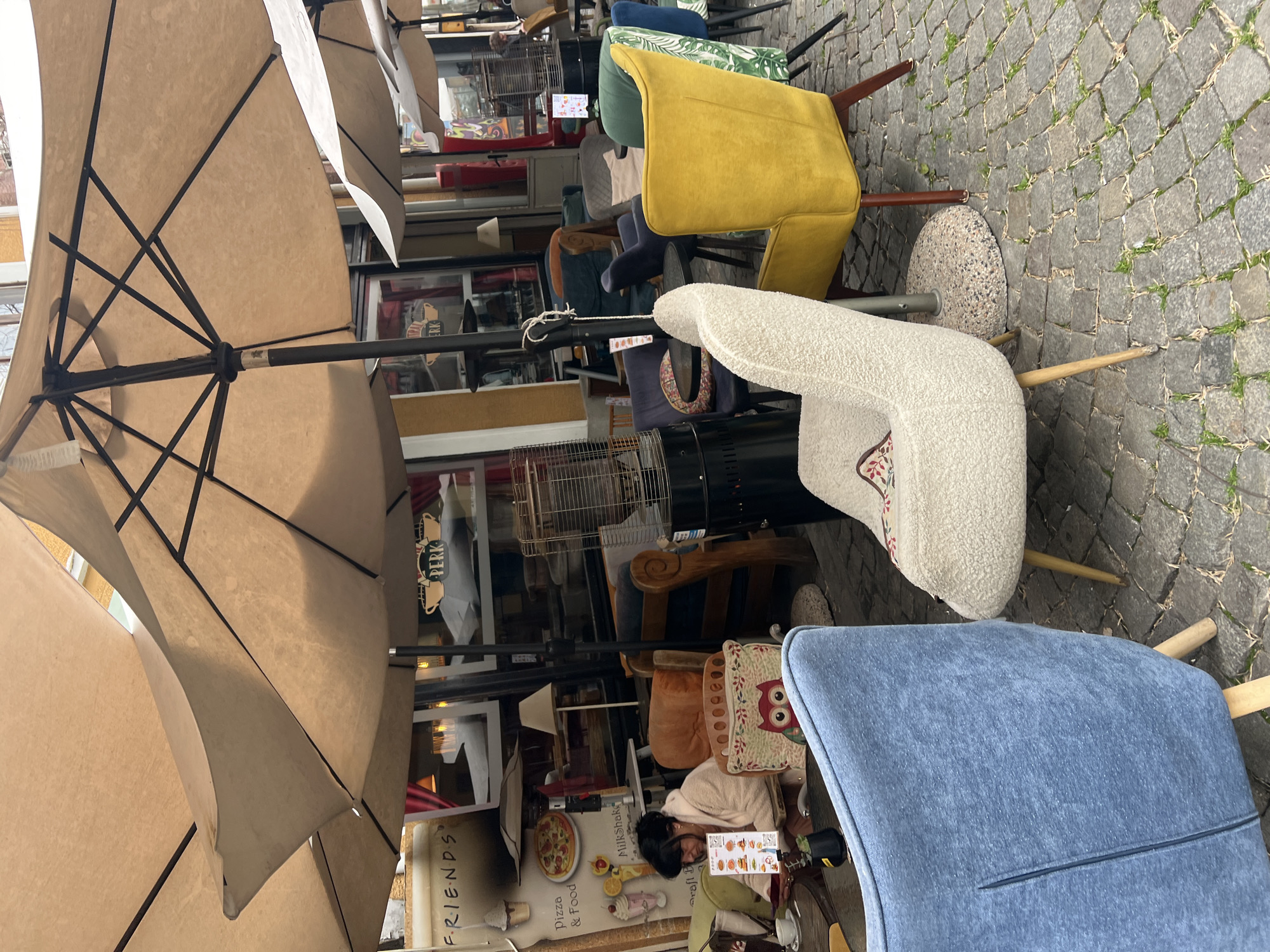 The image depicts an outdoor cafe in Plovdiv, Bulgaria, characterized by cobblestone paving and an eclectic mix of vintage-style chairs arranged seemingly haphazardly. There is a woman with dark hair seated at a small round table near a light blue armchair, seemingly engrossed in a menu or other printed material. The background is cluttered with interior elements visible through a glass storefront, including furniture, signage with the word "FRIENDS," and food advertisements featuring items like pizza and milkshakes. Large beige umbrellas provide shade over the seating area.