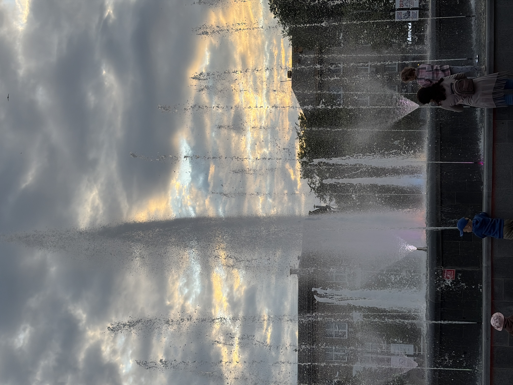 The image shows a public fountain with water jets reaching high into the sky. The sky above is cloudy with patches of golden and blue light, suggesting either sunrise or sunset. In the foreground, a few people are visible, including two children and an adult, seemingly observing the fountain. Buildings with dark roofs and visible windows are in the background. The overall scene appears to be an outdoor public space. The fountain is likely located in the city of Chaikovsky, Russia, as per the provided information.