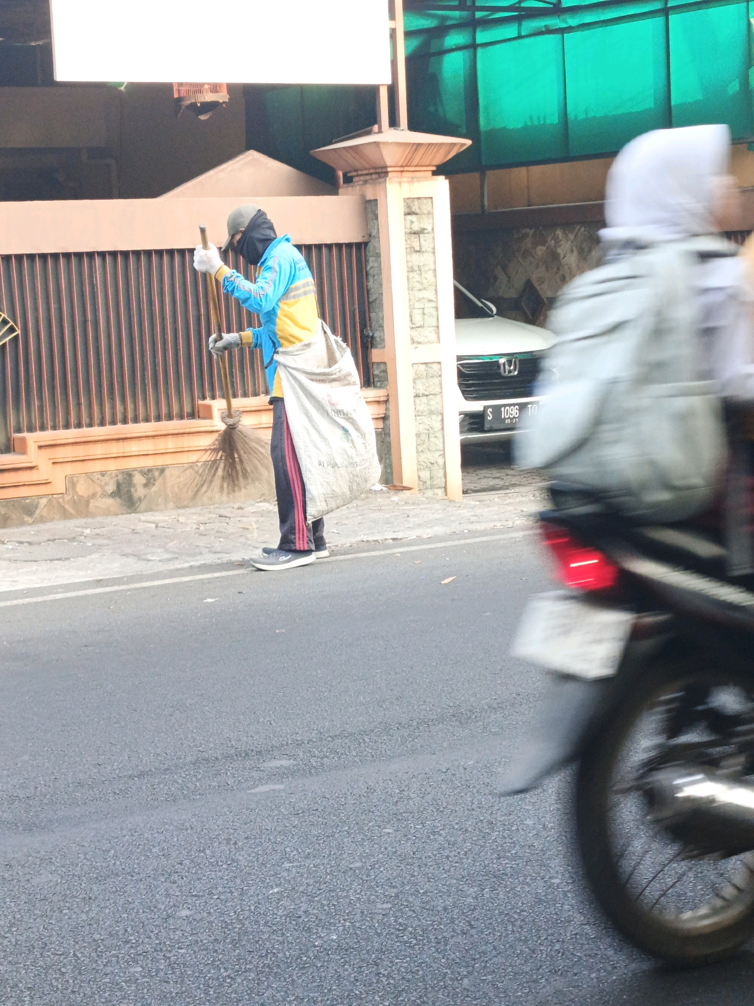 In the city of Mentikan, Indonesia, a street sweeper wearing a blue and yellow uniform, a dark face mask, and a gray cap is visible sweeping the street with a broom. They are carrying a large white sack over their shoulder. A motorbike, with its license plate partially obscured, is in the foreground, its red taillight illuminated. In the background, a white car is parked in front of a building with a brown fence. A person in a light-colored headscarf is blurred in motion on the right side of the image. The scene appears to be a typical street in Indonesia, with elements of urban life and daily activities.