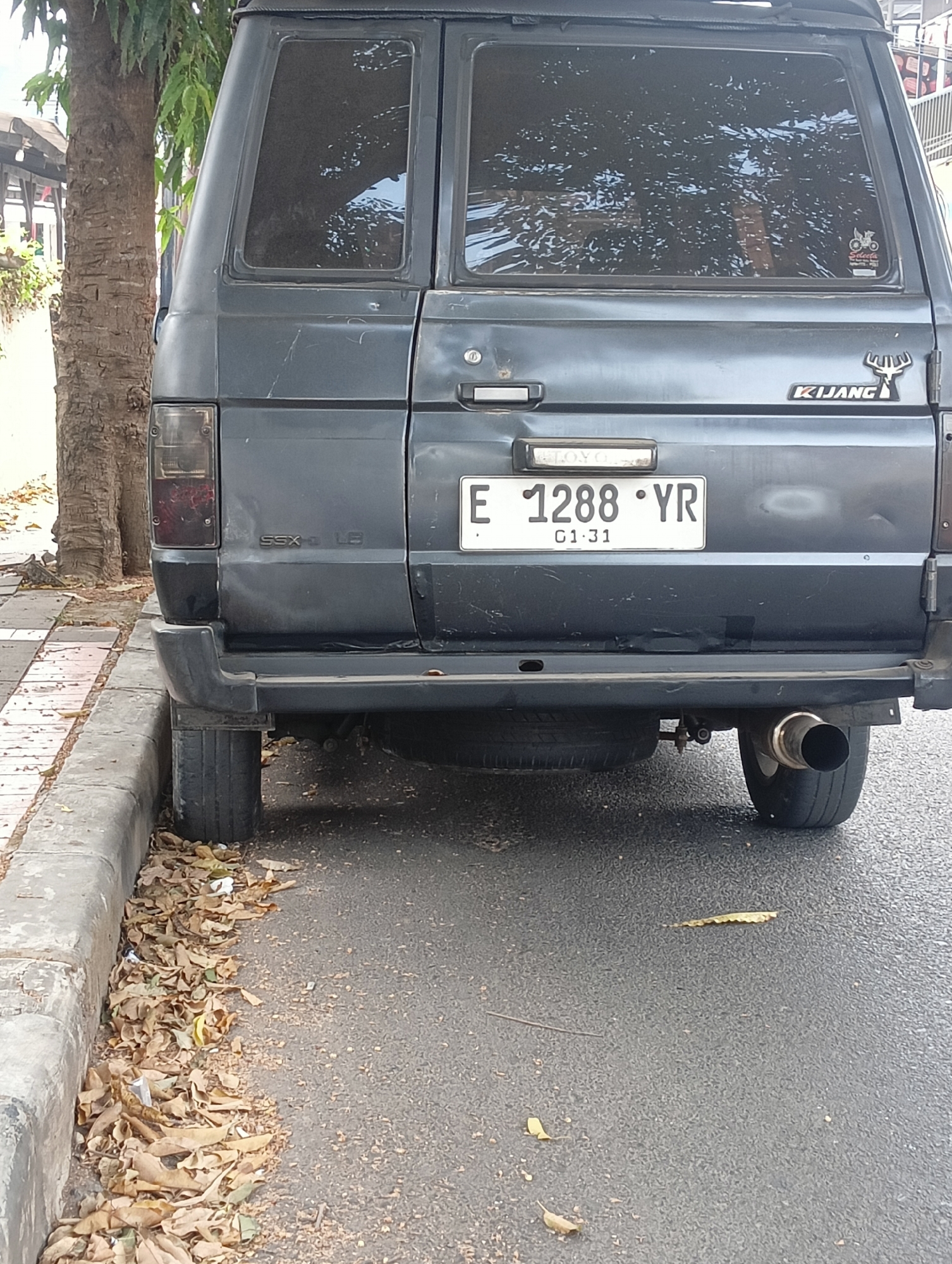 The image shows the rear view of a dark gray van parked on the side of a street in Ciporang, Indonesia. A palm tree is visible to the left of the van. The van has a license plate that reads "E 1288 YR 01-31". There is a spare tire mounted underneath the rear of the van and a silver exhaust pipe extending from the right side. Dry leaves are scattered on the ground next to the curb. The street appears to be asphalt, and the curb is concrete.