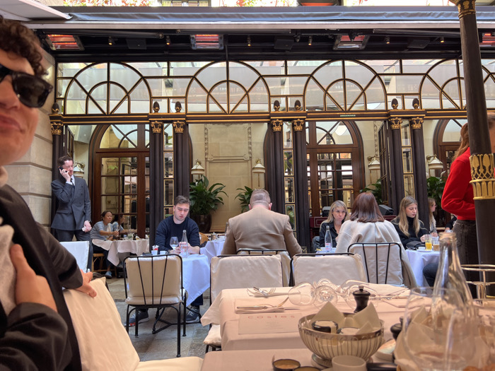 The scene depicts an interior dining area of Hôtel Costes, located in Paris, France, specifically within its restaurant courtyard. The environment is characterized by ornate architecture, featuring dark columns with gilded capitals, large arched windows or doorways with dark frames and gold accents, and light-colored stone walls. Several red-glowing infrared heaters are mounted overhead.
Approximately 10-12 individuals are visible within the space. In the immediate foreground, on the left, a person with dark curly hair, wearing sunglasses and a dark jacket, is partially visible from the chest up, holding a white cloth. In the mid-ground, to the left, a man in a dark grey suit stands next to an arched doorway, engaged in a phone call. To his right, a man in a dark blue long-sleeved shirt is seated at a white-linened table, holding a wine glass, with a bottle of water on the table. Further right, a man in a light brown jacket is seated with his back to the viewer. Adjacent to him, three women are seated at a table, two facing forward and one partially obscured. Additional figures are visible in the background, seated at tables.
All visible tables are covered with white tablecloths and set with white chairs, cutlery, and various glasses. On a table in the foreground, a silver bowl, a salt and pepper grinder, and a clear glass bottle are present. The word "costes" is visible on a placemat or menu item on this table. Potted plants are strategically placed against the architectural elements. The lighting is bright, suggesting natural light from above, supplemented by the artificial glow of the heaters. Patrons are primarily seated, engaged in dining or conversation.