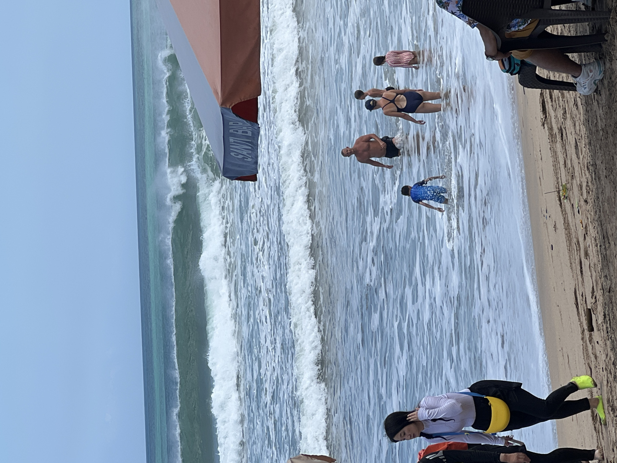 The image depicts a busy beach scene in Kuta, Indonesia. In the foreground, a woman in a white long-sleeved shirt, black leggings, and a yellow fanny pack walks along the sandy shore. Further into the water, several people are enjoying the waves. A man in black shorts stands in the water, while a woman in a dark blue swimsuit, a boy in a blue swimsuit, and a girl in a striped swimsuit are also visible. The ocean is active with moderate waves and a turquoise hue. In the background, a two-toned beach umbrella, one side blue and the other reddish-orange, provides shade, with the word "SANTI BAR" visible on the blue section. The sky is overcast, suggesting a cloudy day. A person sits on a chair in the far right of the image, facing away from the camera.