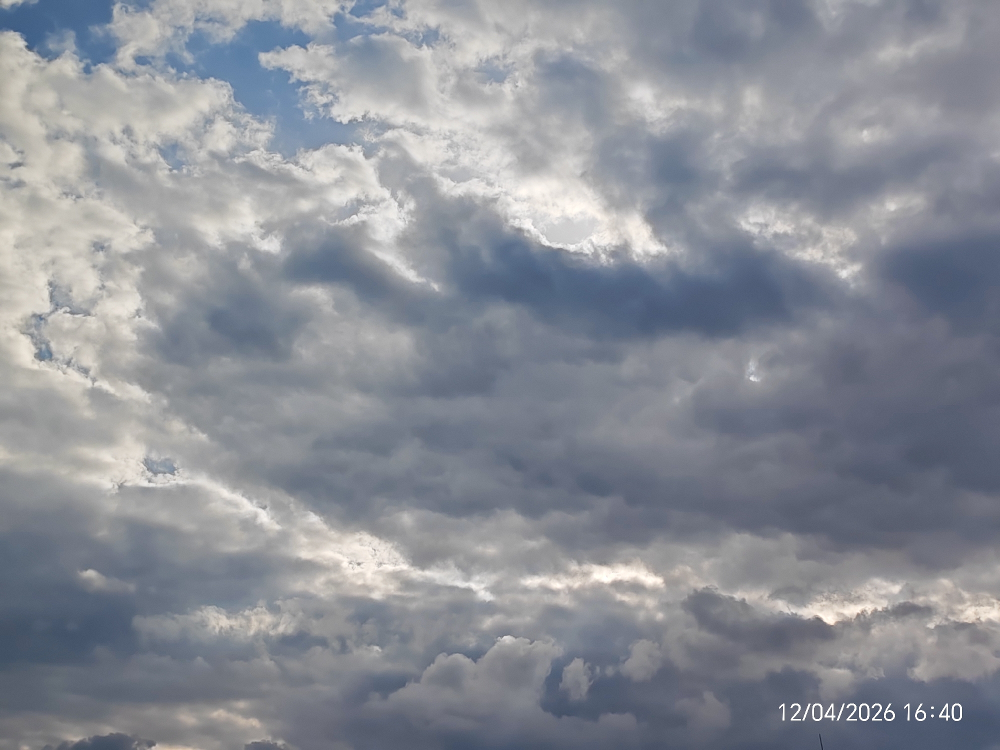 The image shows a sky filled with thick, layered clouds. The clouds vary in color from bright white to dark gray, suggesting varying degrees of sunlight penetration. There are patches of blue sky visible between the clouds. The clouds appear to be dense and create a dramatic, overcast sky. In the bottom right corner, there is a timestamp indicating 12/04/2026 16:40. The location of the city is unknown.