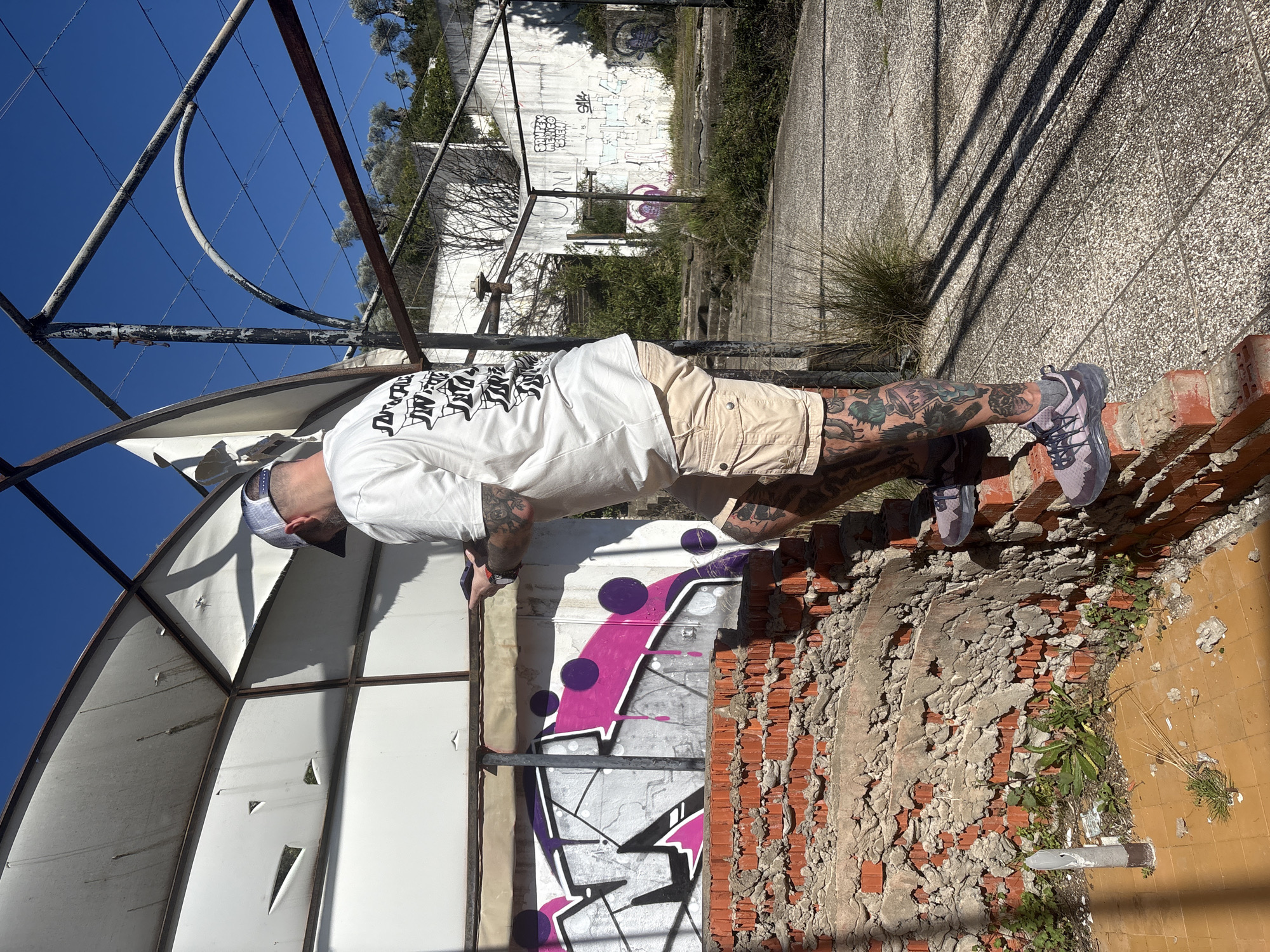 A medium-shot, horizontally oriented image shows a man performing a push-up against an urban ruin. The man wears a white t-shirt with dark lettering, light-colored shorts, a backwards baseball cap, and grey athletic shoes. He is heavily tattooed on both legs and forearms. His hands are pressed against a white wall with purple and black graffiti. His feet are supported by a crumbling red brick wall. In the background is a white wall with graffiti. A corroded, dome-shaped metal structure is to the left of the man. Blue sky is visible in the upper left corner. Debris covers the ground adjacent to the brick wall. This is in the city of Bratica, Braticë, Montenegro.