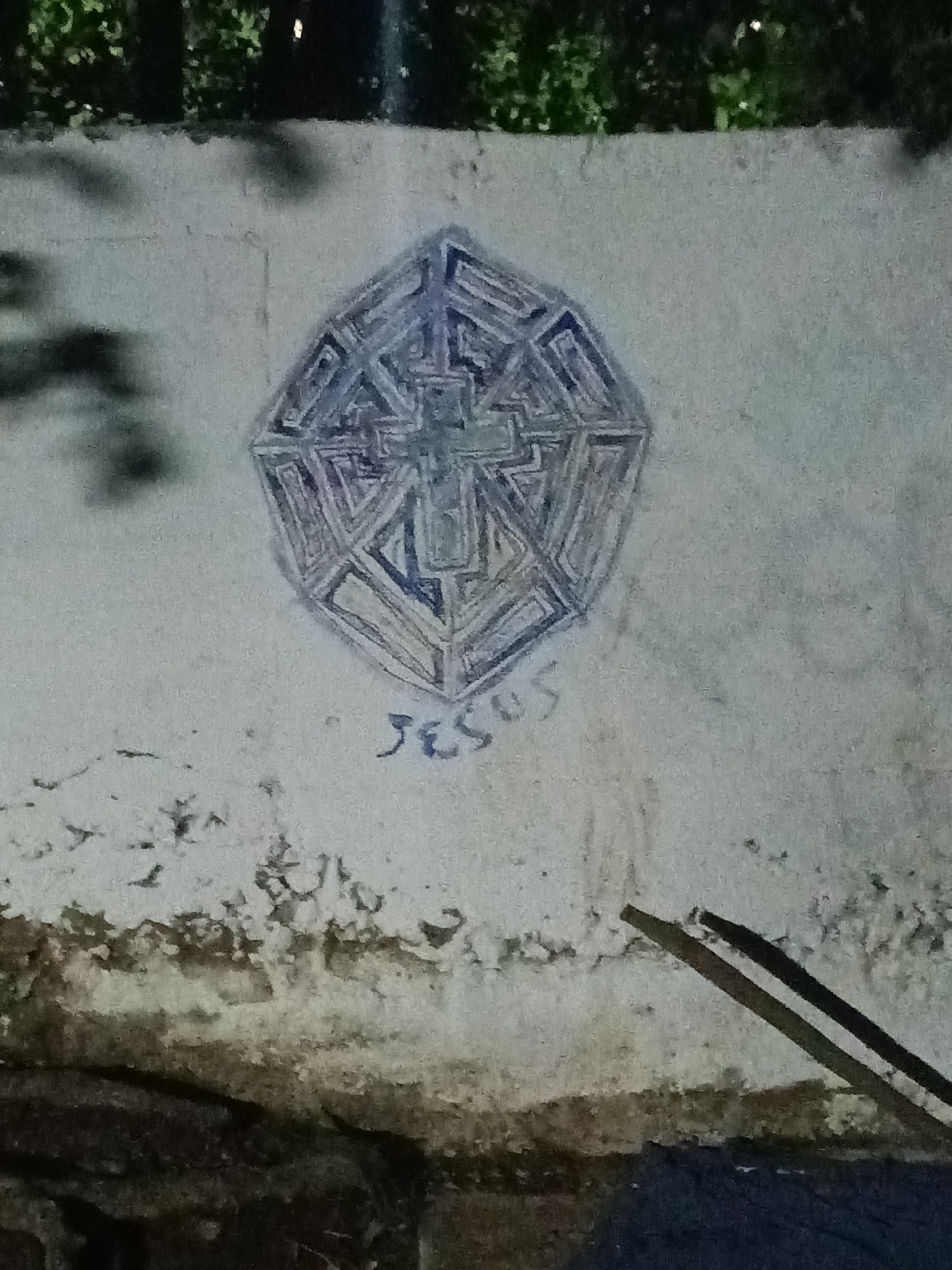 The image depicts a light-colored wall on which graffiti has been drawn. The graffiti consists of a geometric shape, resembling a complex, multi-layered octagon, with a cross at its center. Below the geometric shape, the name "Jesus" is written in a stylized, handwritten font. The wall shows signs of weathering and age, with visible stains and discoloration, especially towards the bottom. In the foreground, partially obscuring the lower right corner of the wall, are two dark, stick-like objects. The background is out of focus, with trees visible at the top of the frame. The location of this image is Tahlequah, United States of America.