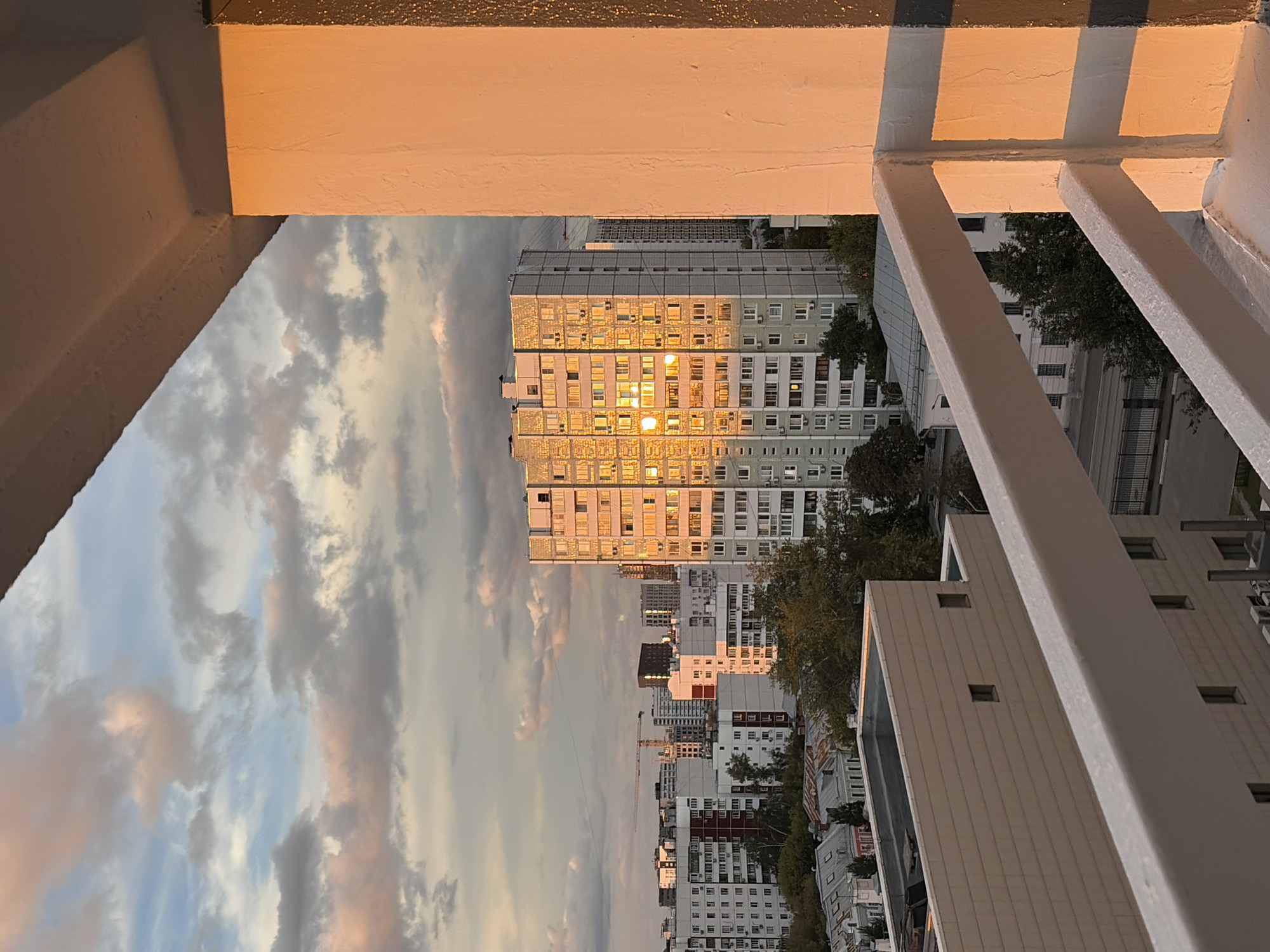 This is a high-angle, full-shot, outdoor photograph of a city skyline at sunset. The perspective is from a balcony, with the right side of the frame dominated by the sunlit exterior of a building, rendered in warm, orange hues. The sky above is filled with a mix of white and grey clouds, tinged with pink and orange by the setting sun.
In the mid-ground, a tall, multi-story apartment building stands out, its facade reflecting the golden light of the sunset, creating bright, dazzling patches. This building is surrounded by other apartment blocks of varying heights and architectural styles, most of which are lighter in color and appear to be residential buildings. There are also glimpses of trees and smaller, older buildings with sloped roofs in the lower portions of the cityscape. A construction crane is visible in the far distance, suggesting ongoing development in the area.
The foreground features a white balcony railing. The scene is devoid of any people or visible action. The overall impression is of a quiet urban evening in Moscow, Russia, with the natural light of sunset dramatically illuminating the built environment.