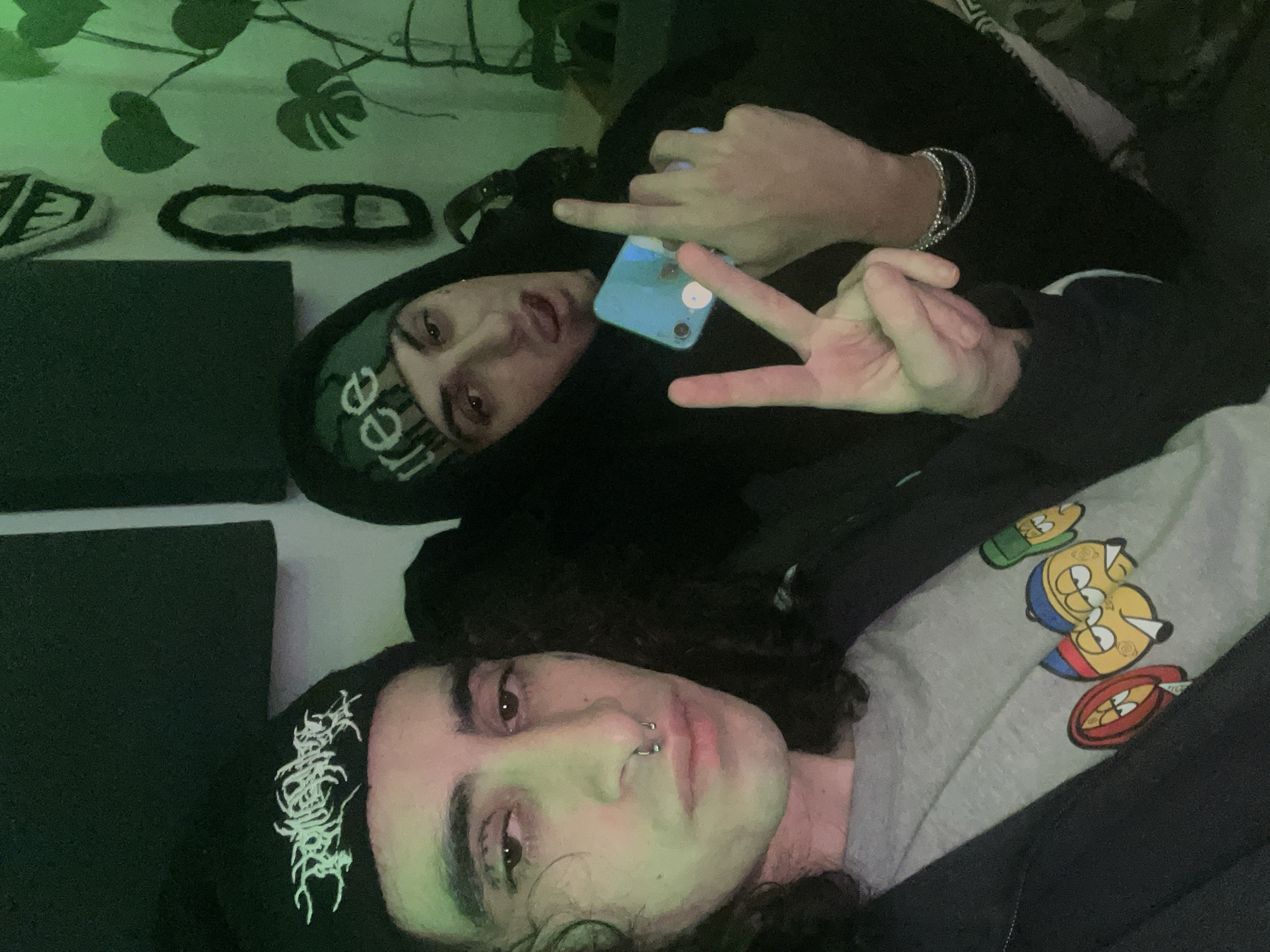 Two people are lying down. The person in the foreground has long curly hair, a nose ring, and is wearing a black shirt with white graphics and a grey sweatshirt with cartoon characters on it. The person in the background is wearing a black hoodie with a green graphic on the forehead and is holding a blue cell phone. The person in the background is also making a peace sign with one hand and raising their middle finger with the other. There are green plants and various objects in the background. The location is not identifiable.