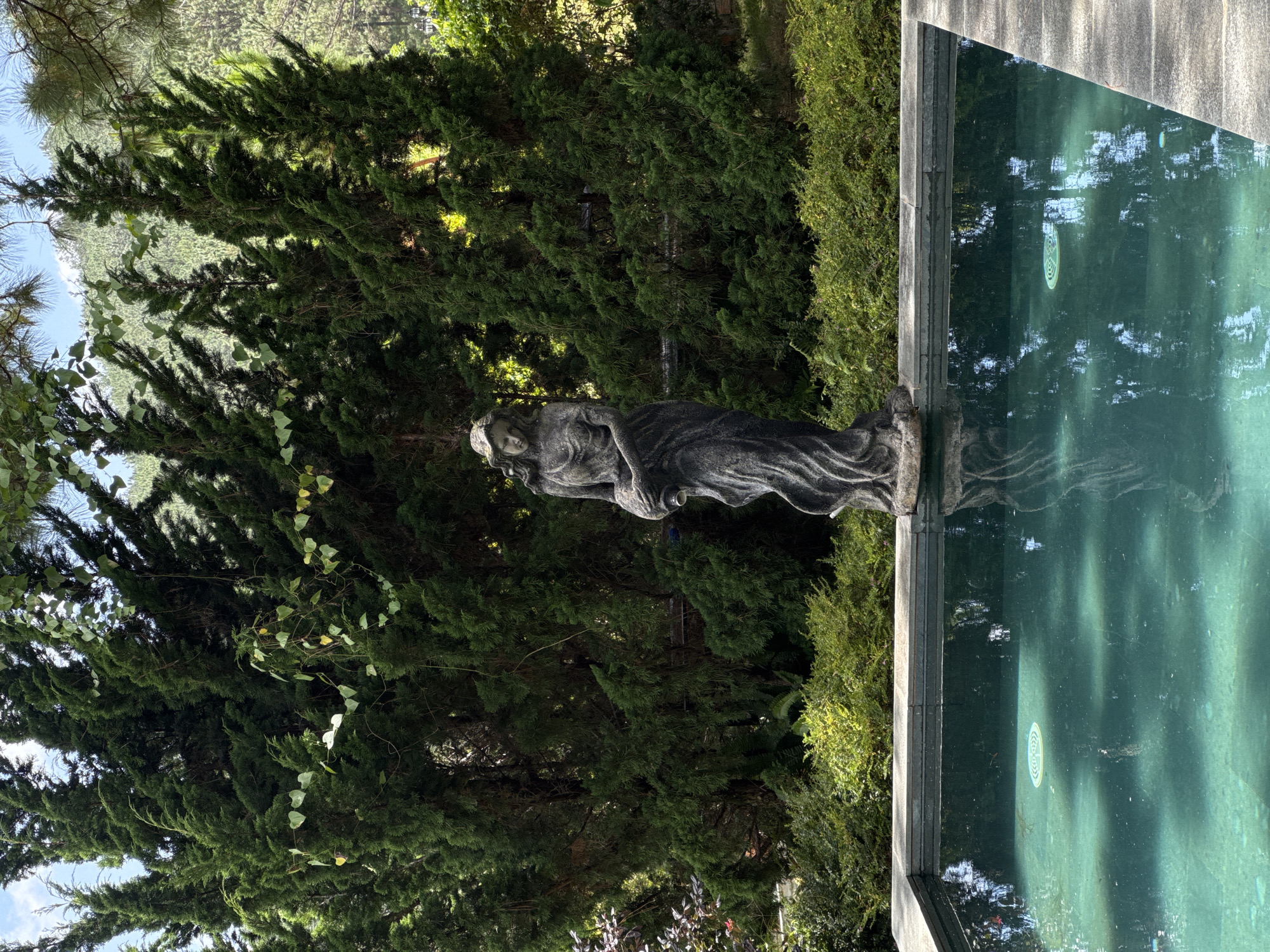 A statue of a draped female figure holding a jug stands in front of a dense forest of cypress trees. The statue is positioned at the edge of a turquoise swimming pool. The pool has a stone coping and reflects the surrounding trees and sky. The background shows a hillside covered in more trees. The location is likely a garden or resort in Da Lat, Vietnam, known for its pine forests and cooler climate.