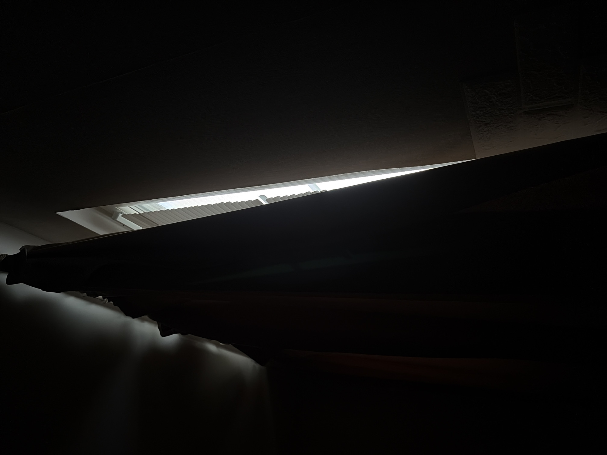 The image presents a high-contrast indoor scene, overwhelmingly dominated by deep shadows and darkness. A striking, bright white light source emanates from a horizontal opening across the upper middle of the frame, likely a window or skylight. This light reveals subtle vertical lines, suggesting the presence of blinds or slats.
Below the light source, a dark, textured, fabric-like object, resembling a curtain or drapery, stretches diagonally from left-to-middle towards the lower right. This object appears to have a wavy, folded texture and plunges the lower and right portions of the image into obscurity. To the far left, a dimly lit, possibly reflective surface is hinted at. In the upper right corner, faint, textured, rectangular patterns are vaguely visible against the dark ceiling or wall, suggesting architectural details.
The image captures a static moment, with no discernible activity or people present. The stark contrast between the intense light and the profound darkness suggests either very early morning or late evening, or a heavily shaded room where external light is the primary illumination. The overall color palette is restricted to blacks, dark greys, and the brilliant white of the light source. No visible text is present. The scene is set in Saint Petersburg, Russia.