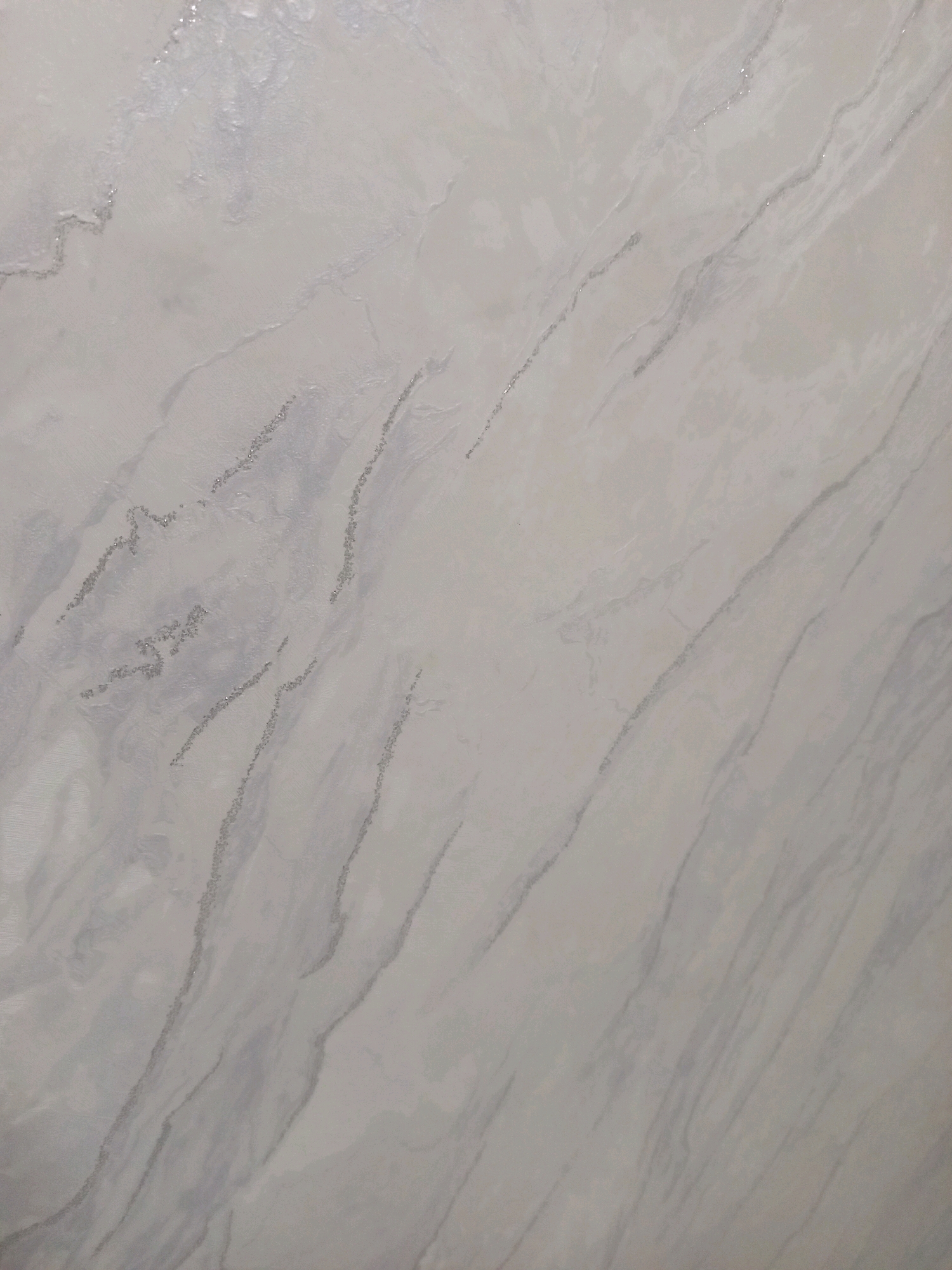 The image is a close-up, overhead shot of a textured, light-colored surface with subtle grey veining and scattered, shimmering particles. The surface appears to be a material like marble or quartz, with a matte finish. The veining runs diagonally across the frame in varying widths and intensities, creating a natural, irregular pattern. The shimmering particles are small and speckled, concentrated along some of the veins, giving the impression of glitter or mica embedded in the material. The lighting is soft and even, casting no harsh shadows. There are no people, animals, or discernable objects in the image other than the surface itself. The overall impression is of a decorative or architectural material. There is no visual information in the image to indicate its location within the city of Jardy-Su, Kyrgyzstan.
