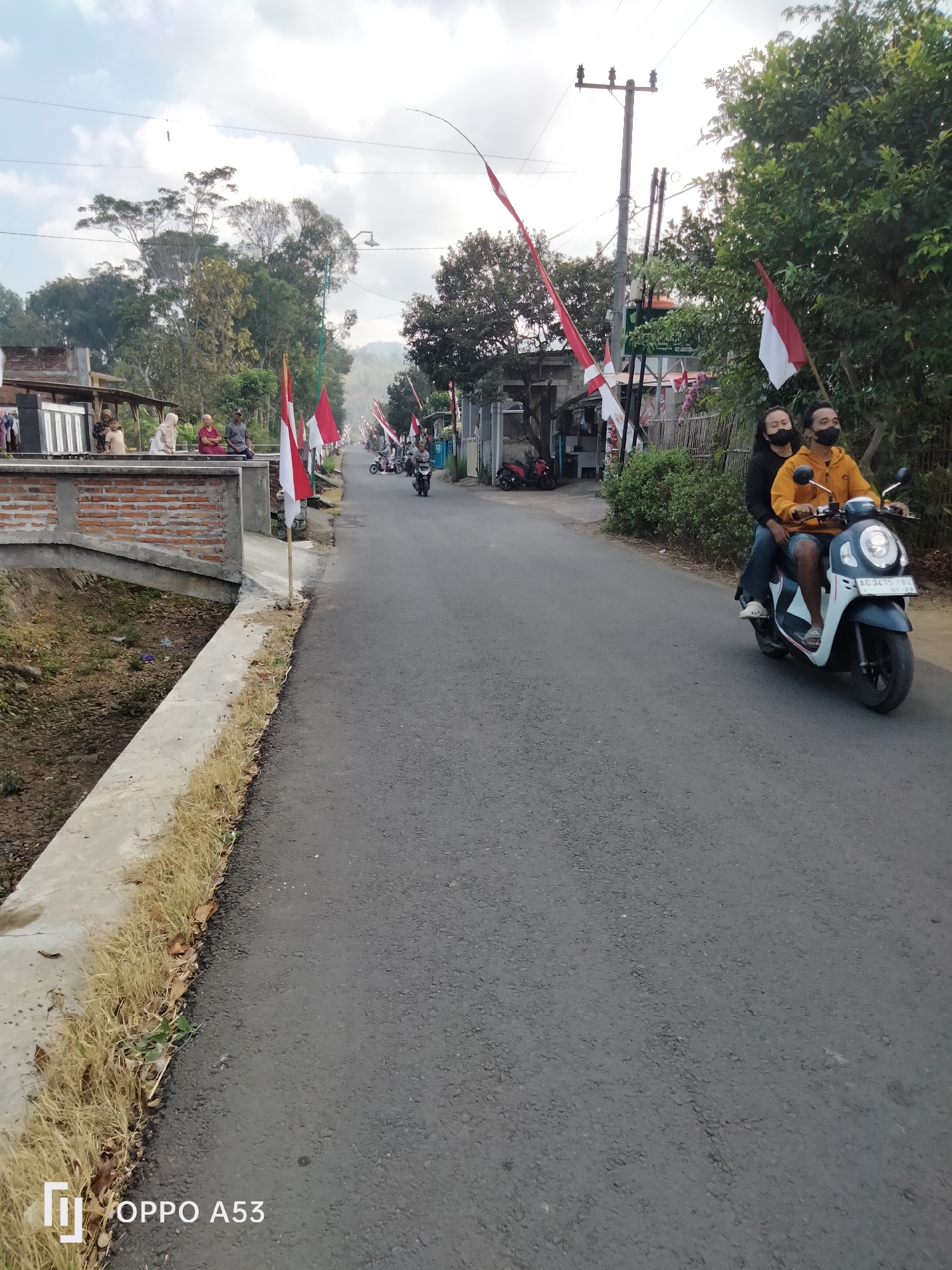 The image shows a street scene in Bendo, Indonesia. In the foreground, two people are riding a scooter, both wearing face masks. The person in front is wearing a yellow hoodie and blue jeans, while the person behind is wearing a black long-sleeved shirt and blue jeans. The scooter is white and blue with license plate AC 3475 YBZ. Red and white Indonesian flags are prominently displayed along the roadside, indicating a celebration or national event.
In the middle ground, several other scooters are visible on the road, with people also wearing face masks. To the left of the road, there is a small brick bridge over a dry ditch. A group of people are seated on a low brick wall near the bridge. Further down the road, small shops or stalls are visible with more flags.
The background consists of lush green trees and vegetation, with a cloudy sky overhead. The overall environment suggests a rural or semi-urban setting in Indonesia, possibly during a period of national observance given the prevalence of the Indonesian flag. The presence of face masks suggests ongoing public health measures.