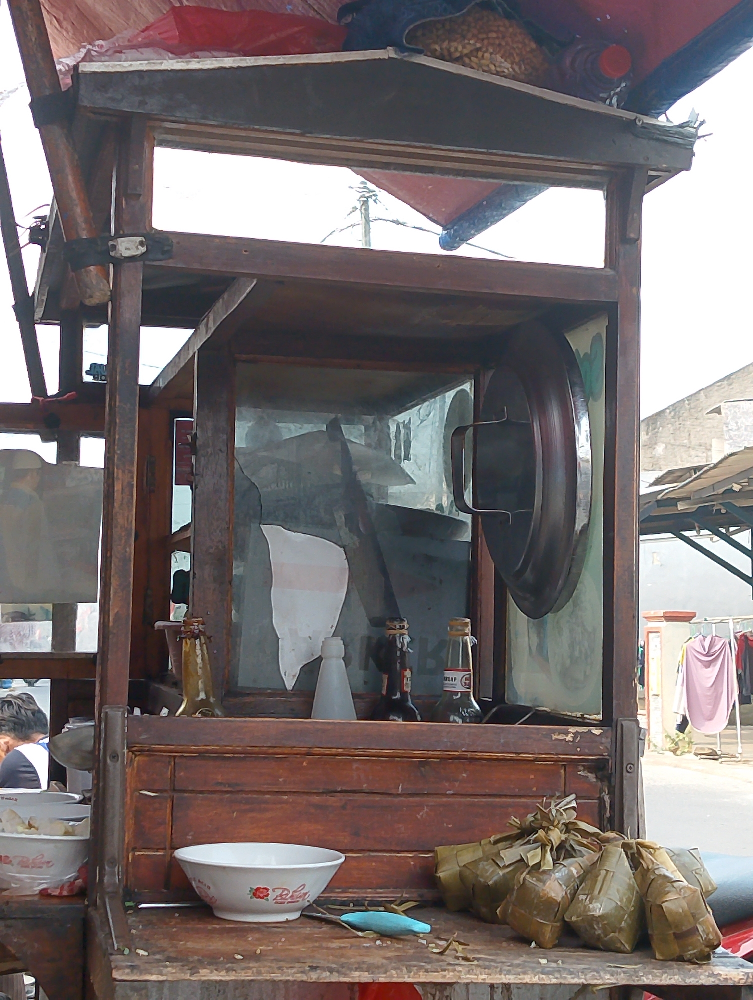 The image captures a street food stall in Karawang, Indonesia. The stall is constructed from dark wood and features a glass display case that appears to be cracked in one section. Inside the case, a large, round metal object with handles is mounted on the back wall. Several bottles, possibly containing sauces or condiments, are visible on a wooden shelf within the stall. In front of the stall, on the wooden counter, are two white bowls, one of which has a floral design and text. Several food items wrapped in leaves, likely rice cakes or similar preparations, are also displayed on the counter. A person's head and shoulders are visible in the upper left portion of the image, suggesting a customer or vendor. The background shows a street scene with buildings and clothing hanging on a rack, indicative of a typical urban or semi-urban environment in Indonesia.