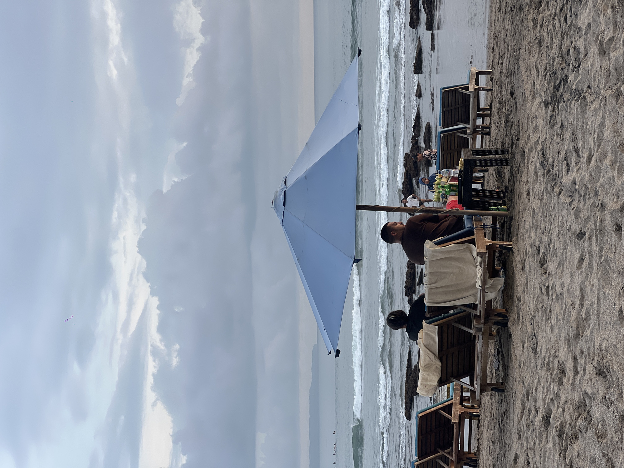 The image shows a beach scene in Canggu, Indonesia. The sky is overcast with heavy clouds. The beach has dark-colored sand, with several lounge chairs and tables arranged under a large, light blue sun umbrella. Two people are visible under the umbrella, one man wearing a brown shirt and jeans, the other person, next to him, is wearing a dark-colored shirt. Nearby, a small table displays what appears to be fruit. Waves are crashing on the shore near rock formations. In the distance, surfers are visible in the ocean. The image is rotated 90 degrees clockwise.