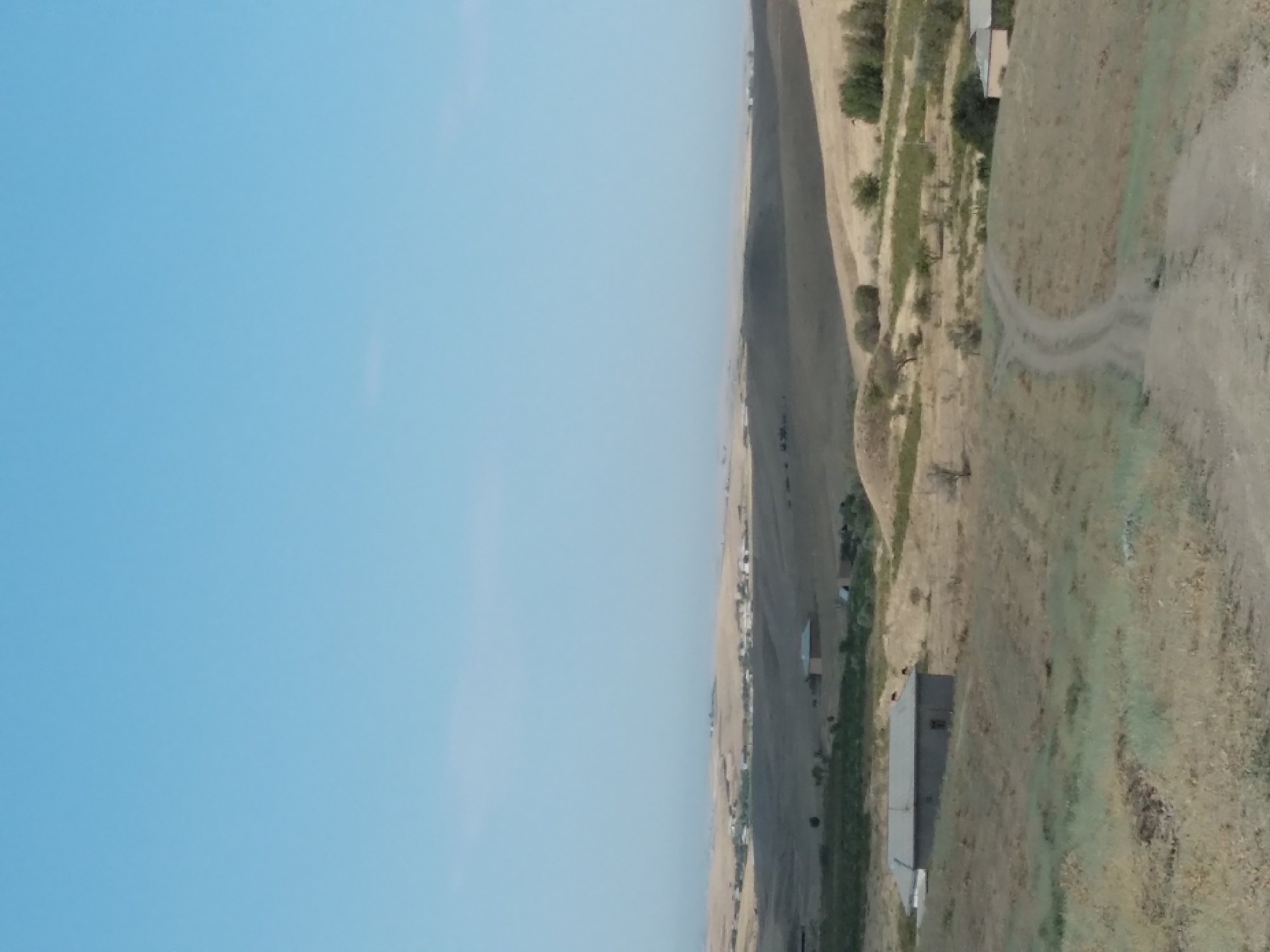 An outdoor, full-frame, eye-level shot captures a landscape with rolling hills under a clear blue sky. In the foreground, a dirt road curves upwards and to the right. To the left of the road is a small, single-story building with a gray roof and light-colored walls. Further into the mid-ground, more single-story buildings with dark roofs are visible amidst dry, grassy terrain. In the far distance, a cluster of buildings suggests a small village or settlement on a distant hill. The overall environment appears arid and rural, likely in or around Aktepa, Uzbekistan, given the dry, steppe-like vegetation and scattered human settlements typical of the region.