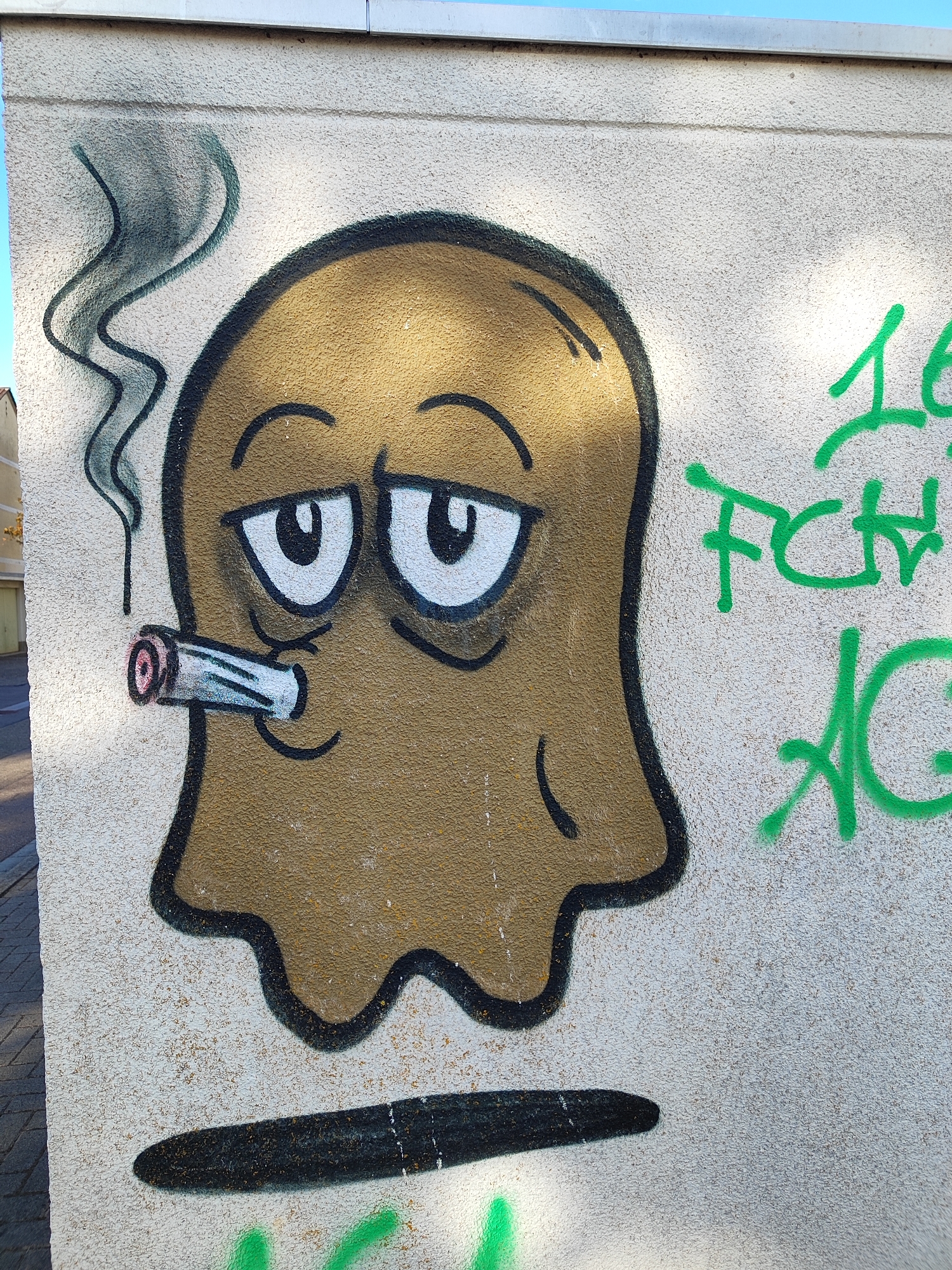 The image shows graffiti on the side of a building in Offenburg, Germany. The graffiti features a cartoonish ghost character with a brown body, black outlines, and large, stylized eyes. The ghost is depicted smoking a cigarette, with gray smoke rising from it. To the right of the ghost, there is green spray paint graffiti that appears to be text, possibly initials or a tag. The building itself has a textured, light-colored facade, and a section of the sky is visible at the top of the frame. The bottom of the image shows a shadowy, dark shape painted on the wall below the ghost, and some green spray paint markings on the ground level. The overall scene is a close-up of street art.