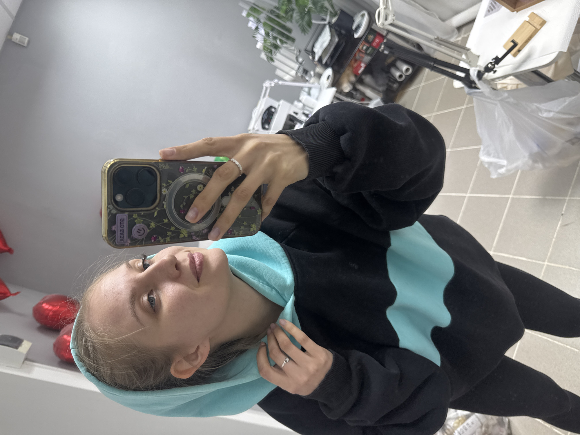 A young woman is depicted taking a selfie indoors, holding a smartphone with a floral-patterned case and a circular phone grip in her right hand. She is wearing a black hoodie featuring a distinct light blue wavy pattern on the front and a matching light blue hood, which she is adjusting with her left hand. Her light-colored hair is parted on the side, and she has a calm, slightly smiling expression. She wears a white, wavy-patterned ring on her right index finger and another ring, possibly with text, on her left ring finger.
The setting appears to be a professional indoor space, possibly a beauty salon or studio, identifiable by the white tiled floor and various pieces of equipment in the background. These include adjustable metal arms or stands, shelves, and what might be a workstation. To the left, several red heart-shaped balloons are visible. On the right, a white plastic bag and a wooden phone stand rest on a surface. Artificial plants or greenery are also subtly present in the background.
The activity is clearly a self-portrait taken with the phone's camera, with the woman's gaze directed towards the phone. The time of day is not evident, but the scene is brightly illuminated by indoor lighting. Text visible on her phone case, appearing reversed due to the reflection, reads "310 В3УЛ" or "ЛУЗВ О1Е" when oriented correctly. The location context for this image is Izhevsk, Russia.