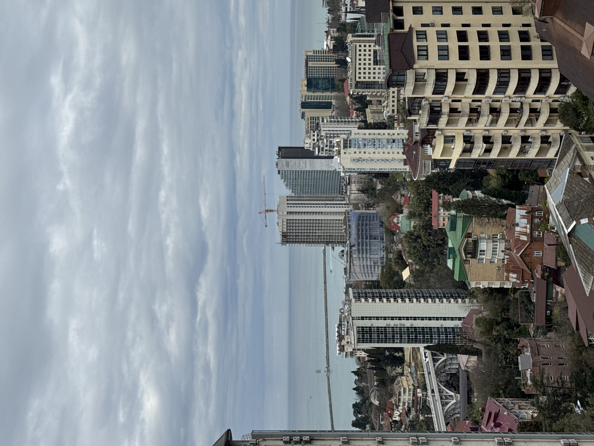 The image is a cityscape in Sochi, Russia, taken from an elevated perspective. It is overcast with a view of the Black Sea. There are many buildings, including large hotels and apartments. There is one construction crane visible. Trees are interspersed between the buildings. A pier extends into the sea.