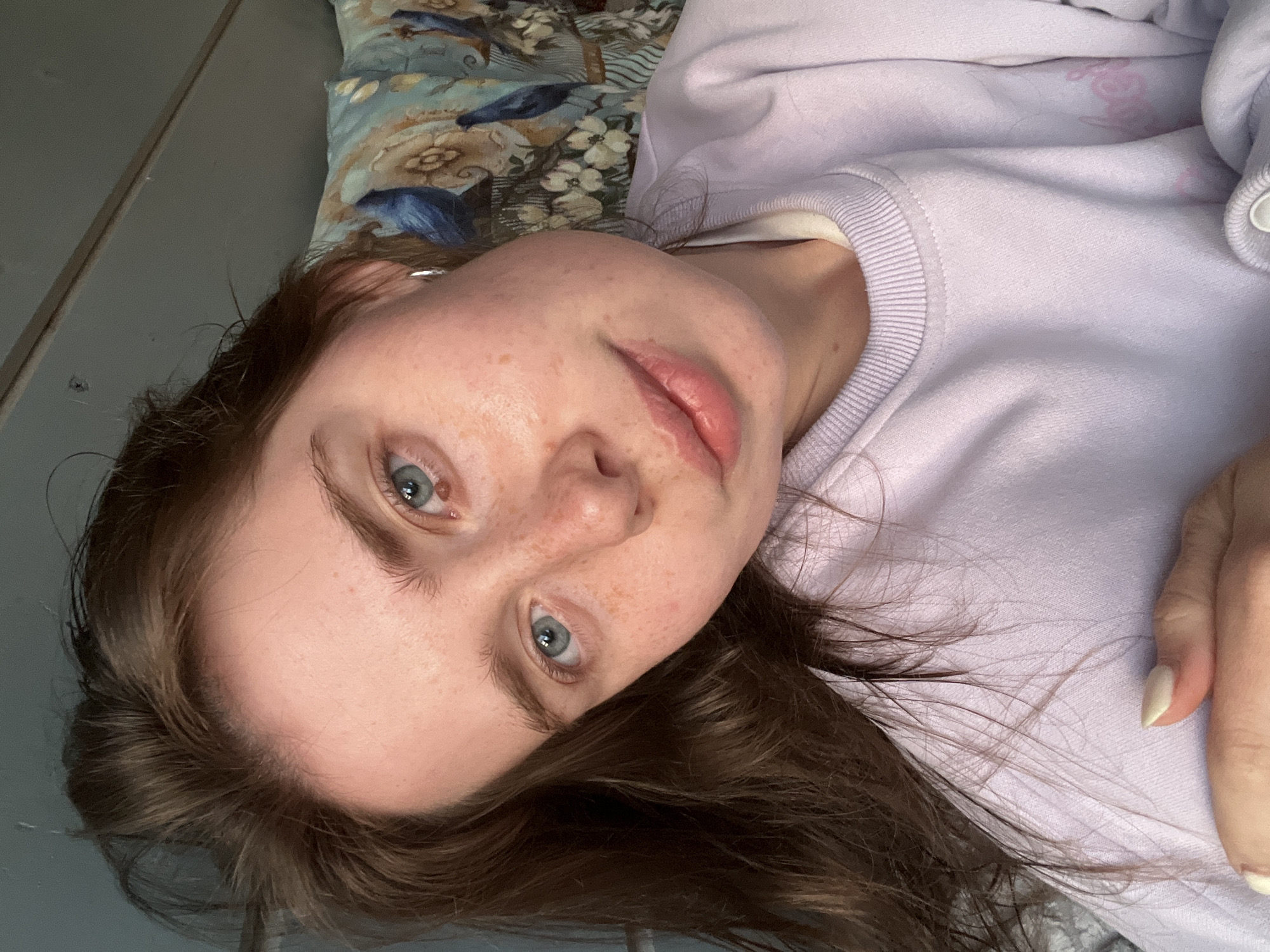 A young woman is lying on a bed. She has fair skin, light blue eyes, and brown hair. Her face is freckled. She is wearing a light purple sweatshirt. The bed has a patterned pillow. The location is not evident in the image, and there is no discernible information to determine if it is in Chaikovsky, Russia, or any other location.