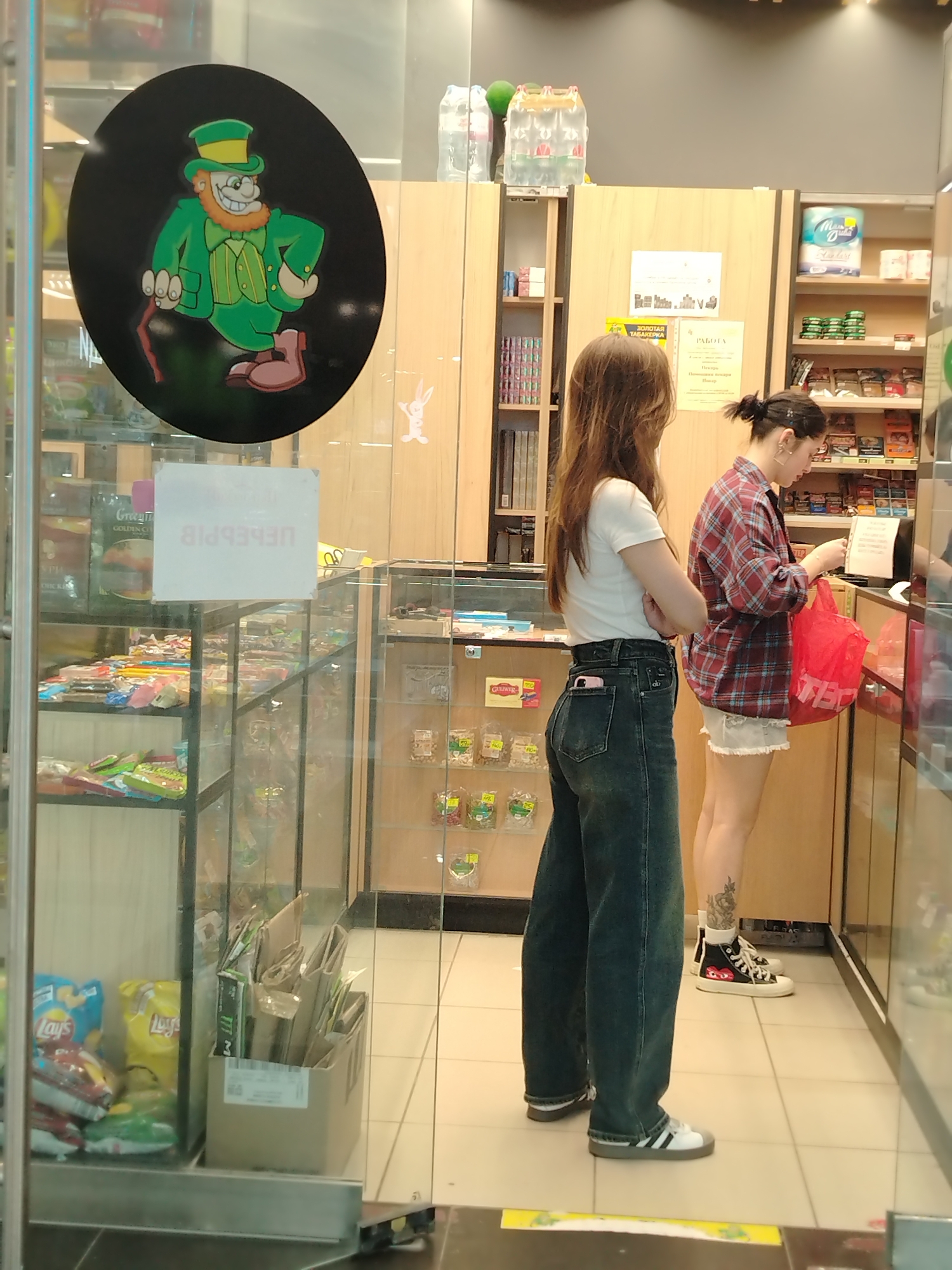 In Izhevsk, Russia, two young women are captured in a medium shot inside what appears to be a convenience store or kiosk. The woman on the left, with long brown hair, is wearing a white t-shirt and dark, wide-leg jeans. Her arms are crossed, and a pink smartphone is visible in her back pocket. She stands facing right. The woman on the right, with dark hair pulled back in a bun, wears a red plaid shirt and light-colored shorts. She is engaged in a transaction, holding a red plastic bag and reaching towards the counter. She is wearing black high-top sneakers with a distinctive design on the side and has a tattoo on her left calf.
The environment is a brightly lit retail space with glass display cases filled with merchandise. To the left, a large circular sticker on the glass depicts a cartoon leprechaun in a green suit and top hat. To the right, shelves are stocked with various items, including packaged goods, drinks, and rolls of toilet paper. The floor is tiled. A white sign with Cyrillic lettering is affixed to the glass in front of the woman on the left. The overall scene suggests a typical commercial exchange within a small store setting.