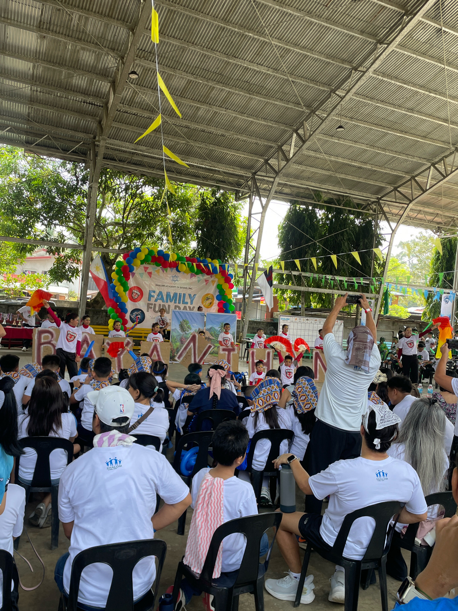 This is a wide-angle, eye-level, full shot of an outdoor event at the Shalom Academy of Davao, Inc. in Davao City, Philippines. The event, a "Family Fun Day," appears to be taking place under a covered, open-air structure with a corrugated metal roof supported by metal beams. The ground is paved.
In the background, a stage or performance area is set up with a banner that reads "SHALOM ACADEMY OF DAVAO, INC. FAMILY FUN DAY." The banner is decorated with balloons and flags. A Philippine flag is prominently displayed to the left of the banner. Children in matching white t-shirts with blue graphics are performing on stage, some holding fans and colorful flags. Several other children and adults are seated in rows of black chairs, facing the stage. Many of these seated individuals are also wearing white t-shirts with the same blue graphics, suggesting they are participants or attendees of the event.
A man in the foreground, facing away from the camera, is holding up two smartphones to record the performance. He is wearing a white t-shirt, black shorts, and a headscarf. Other people are also visible in the foreground, some seated and some standing. Lush green trees and parts of buildings are visible beyond the covered structure, indicating a tropical environment. The lighting suggests it is daytime, with some overcast conditions or shade from the roof. The overall atmosphere seems to be festive and community-oriented.